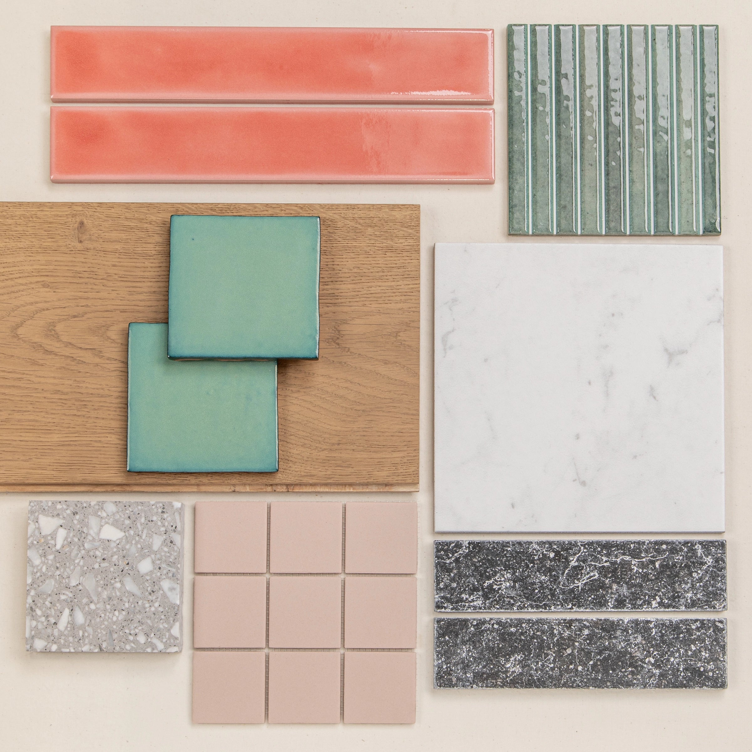 Flat lay of green textured rectangular tiles, coral pink ceramic tiles, wood sample, marble-look tile, terrazzo, beige square tiles, and dark stone tiles on neutral background