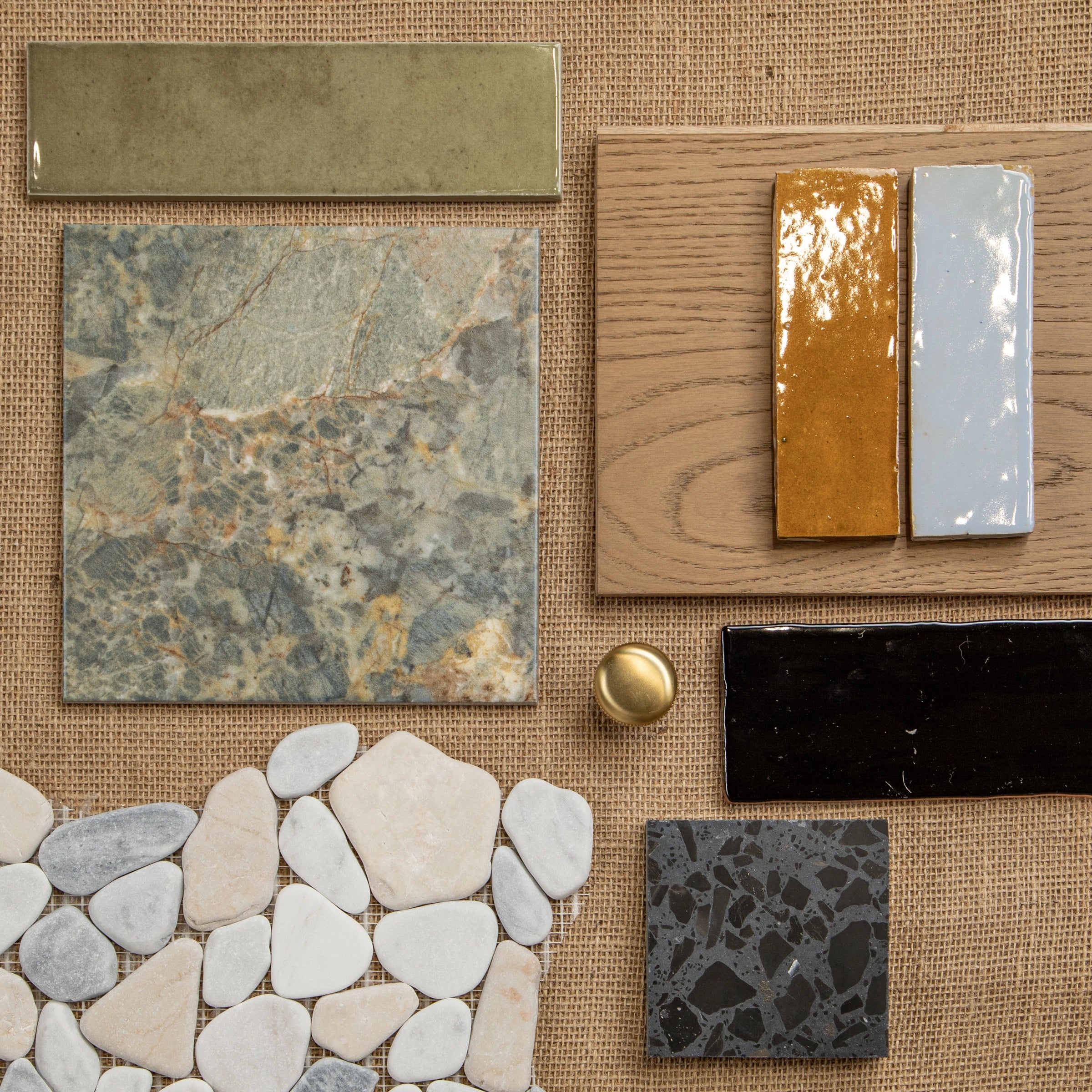 Varied ceramic tiles, pebble shapes, wood plank, and brass knob samples arranged on woven natural fabric background