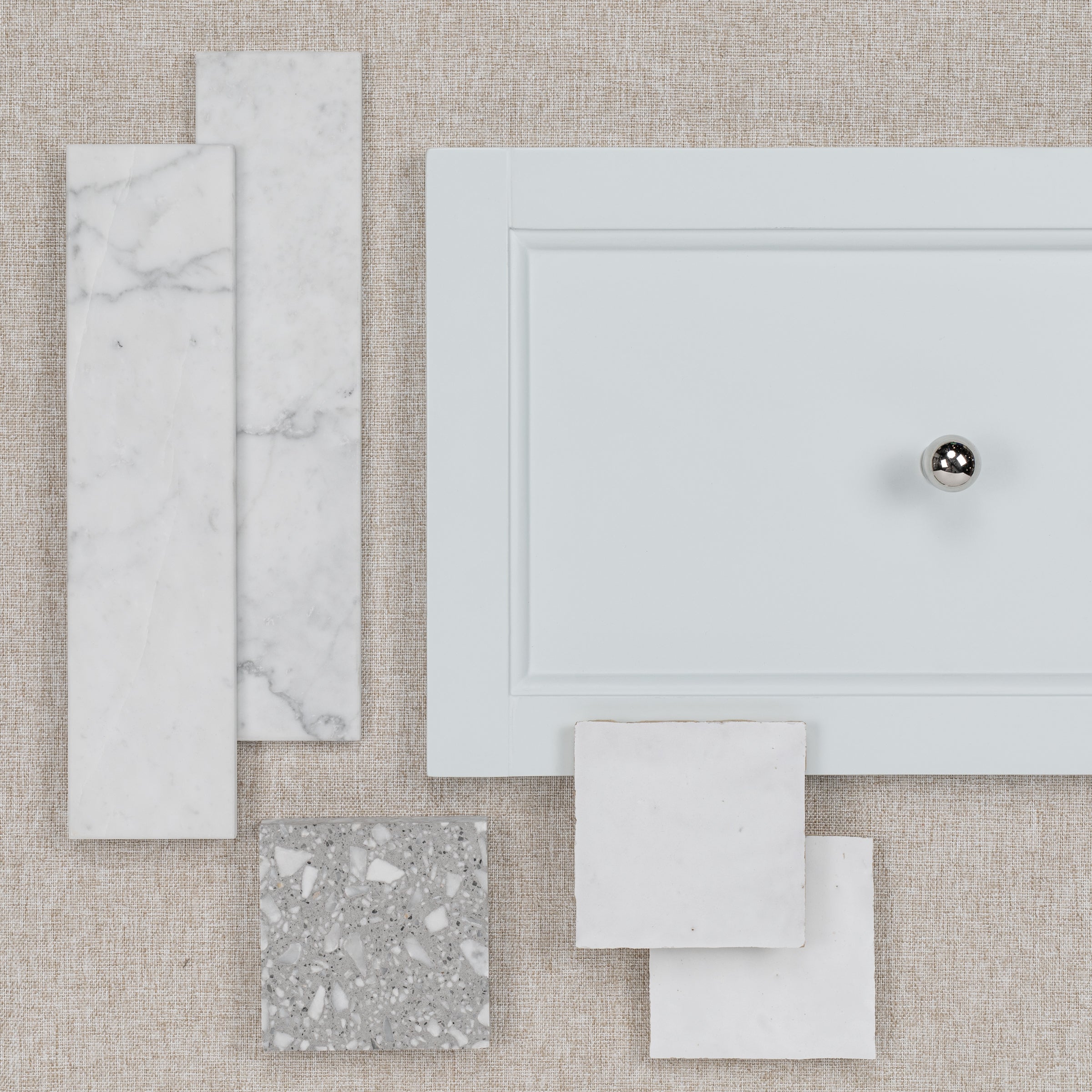 Flat lay of marble tile samples and a light gray cabinet door with a shiny metal knob on a textured surface