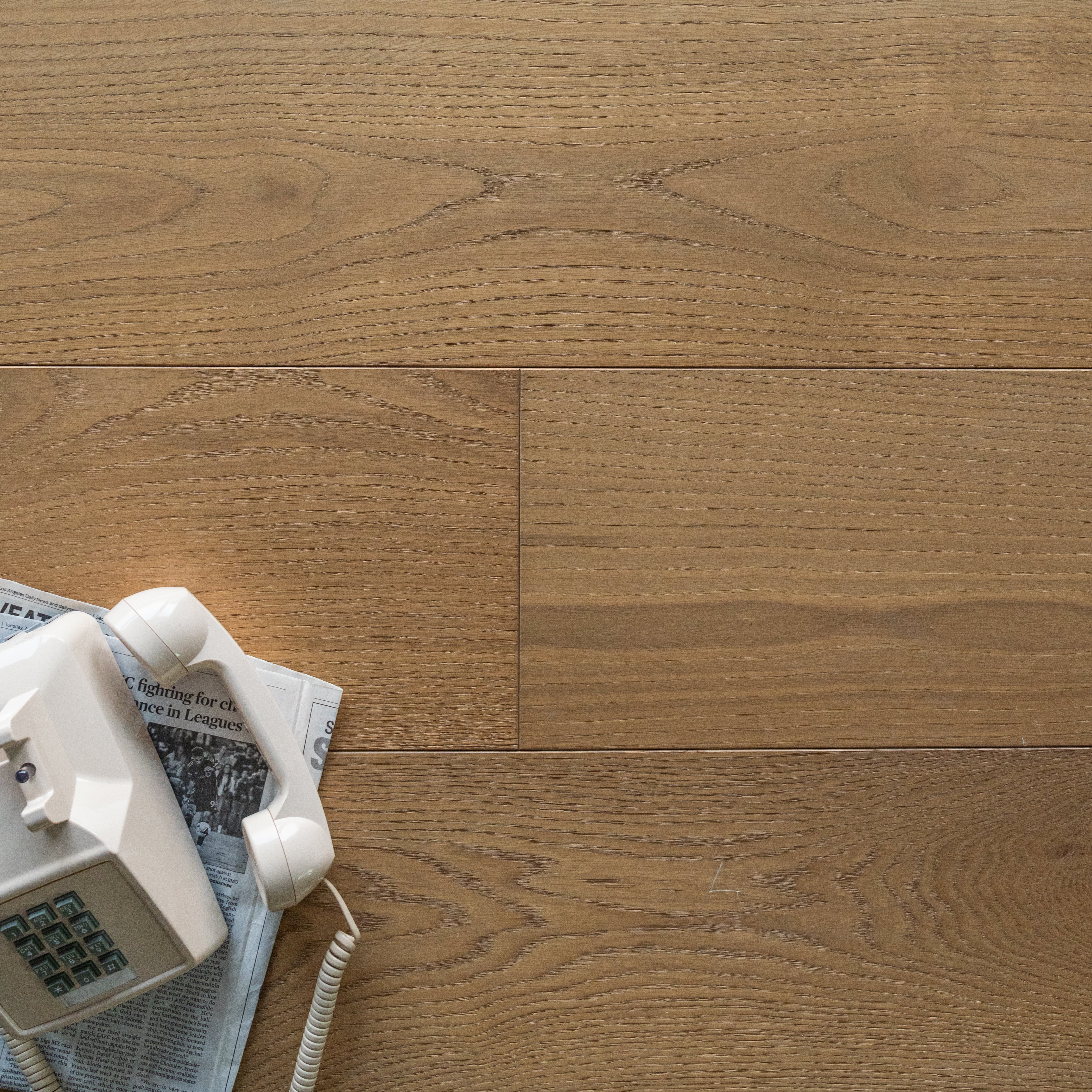 Light brown engineered wood flooring planks with subtle grain pattern and a vintage white corded telephone on newspaper corner