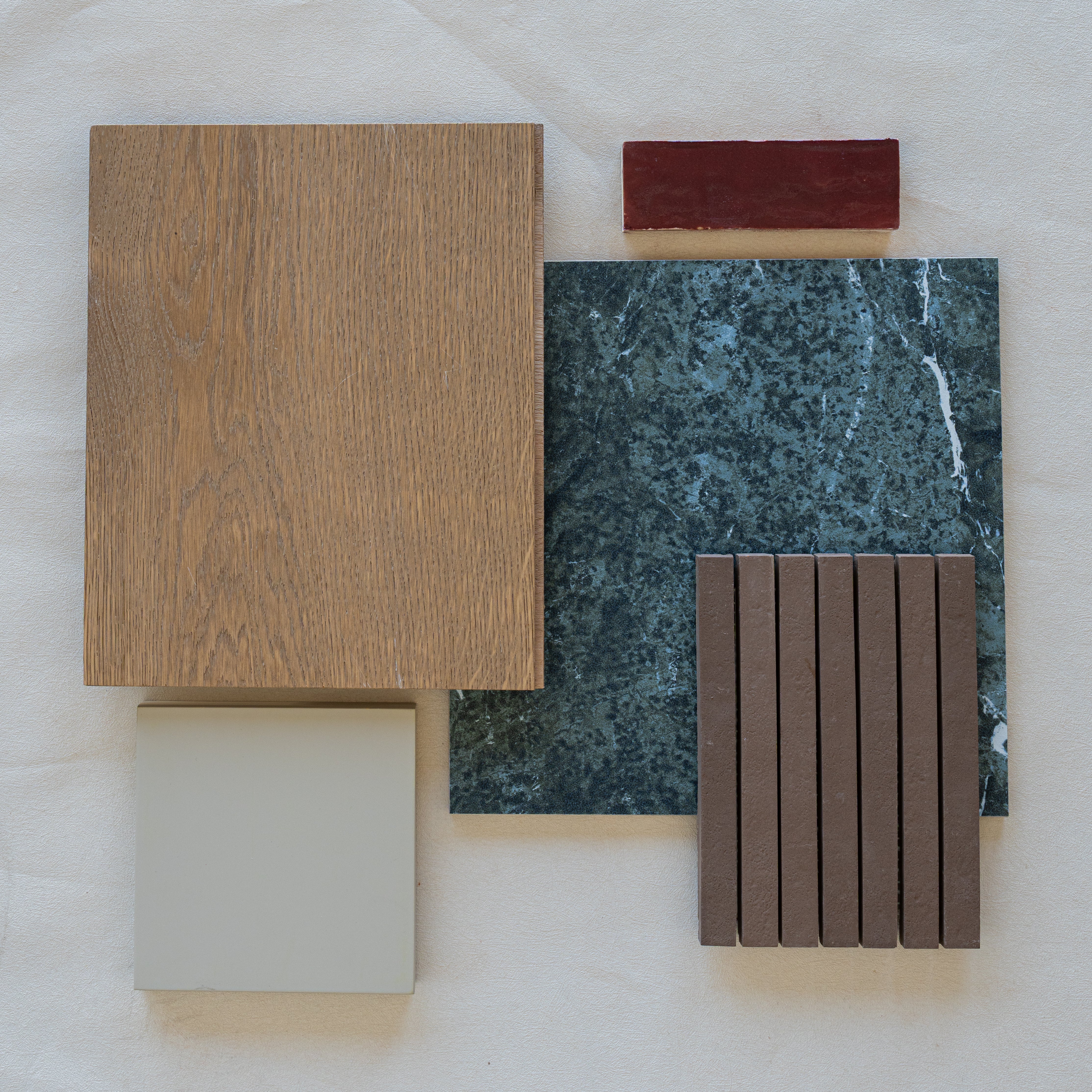 Flat lay of oak wood sample, green marble tile, beige square tile, dark narrow strips, and small red rectangular block on fabric background
