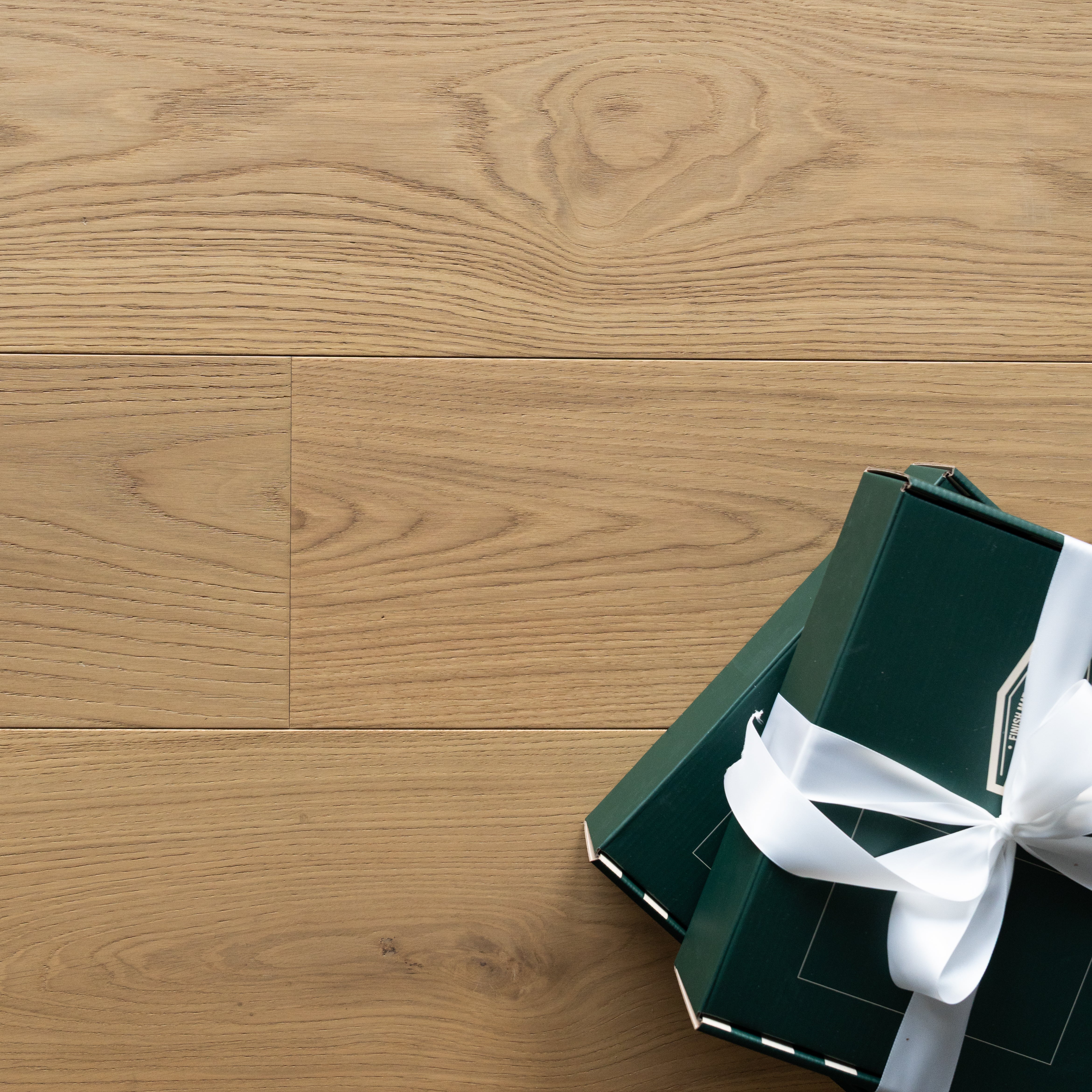 Light oak engineered wood flooring with subtle wood grain pattern and two dark green gift boxes tied with white ribbon in corner