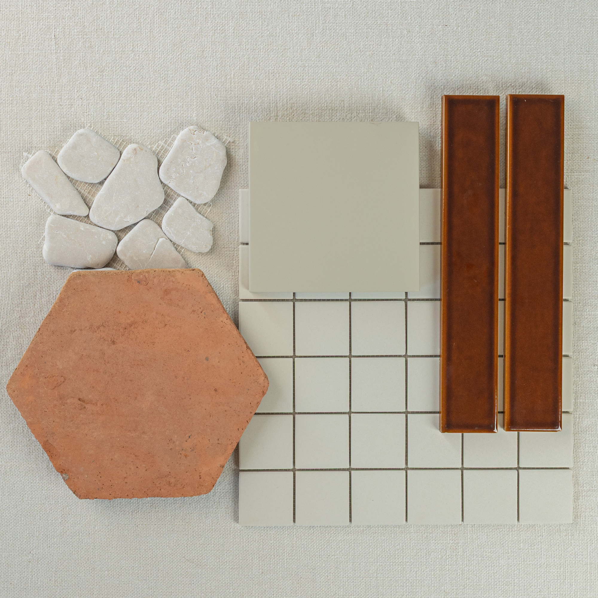 Flat lay of terracotta hex tile, white stone pebbles, beige ceramic squares, and brown glossy rectangular tiles on neutral fabric