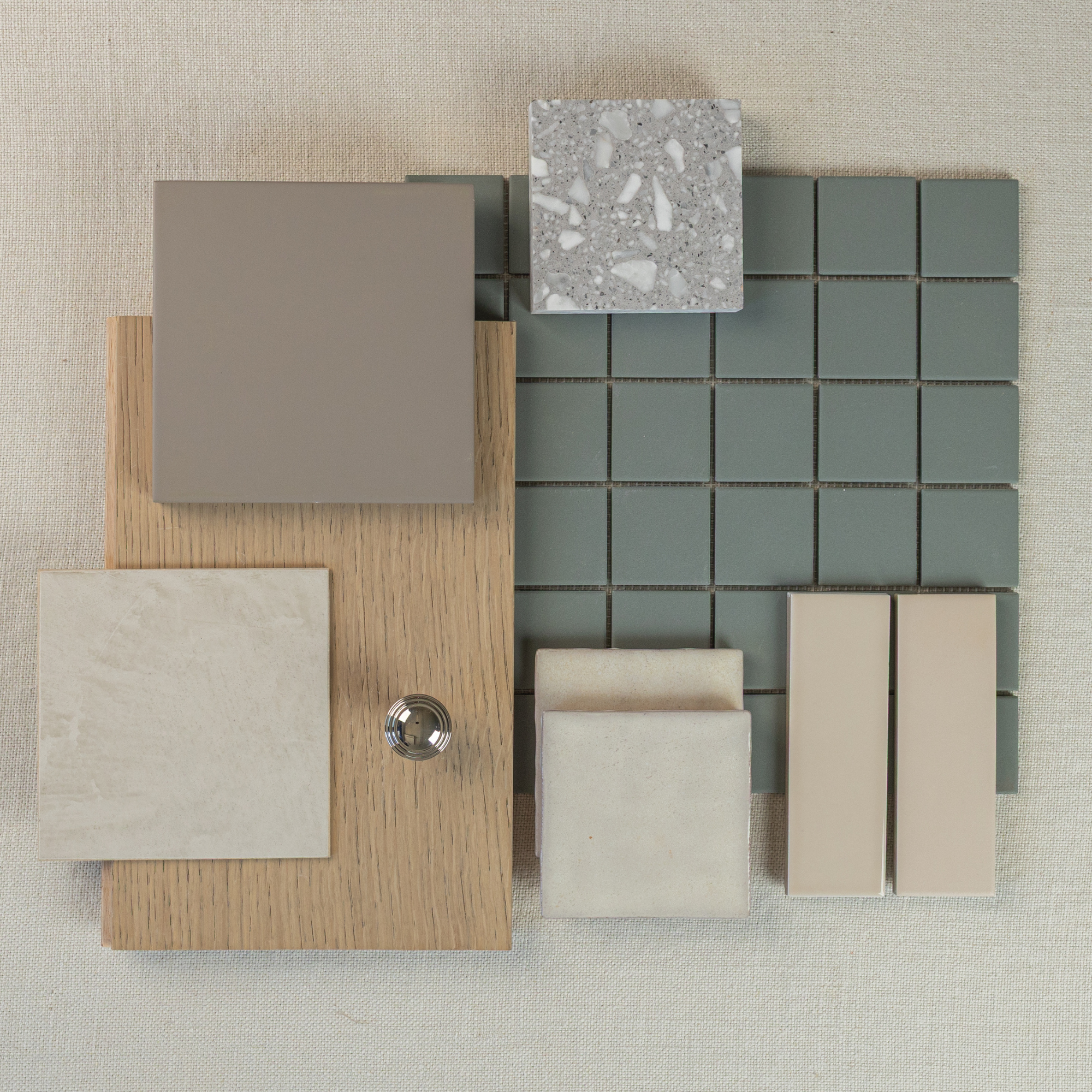 Arrangement of muted green mosaic tiles, beige ceramic tiles, stone sample, wood swatch, and a chrome knob on neutral fabric
