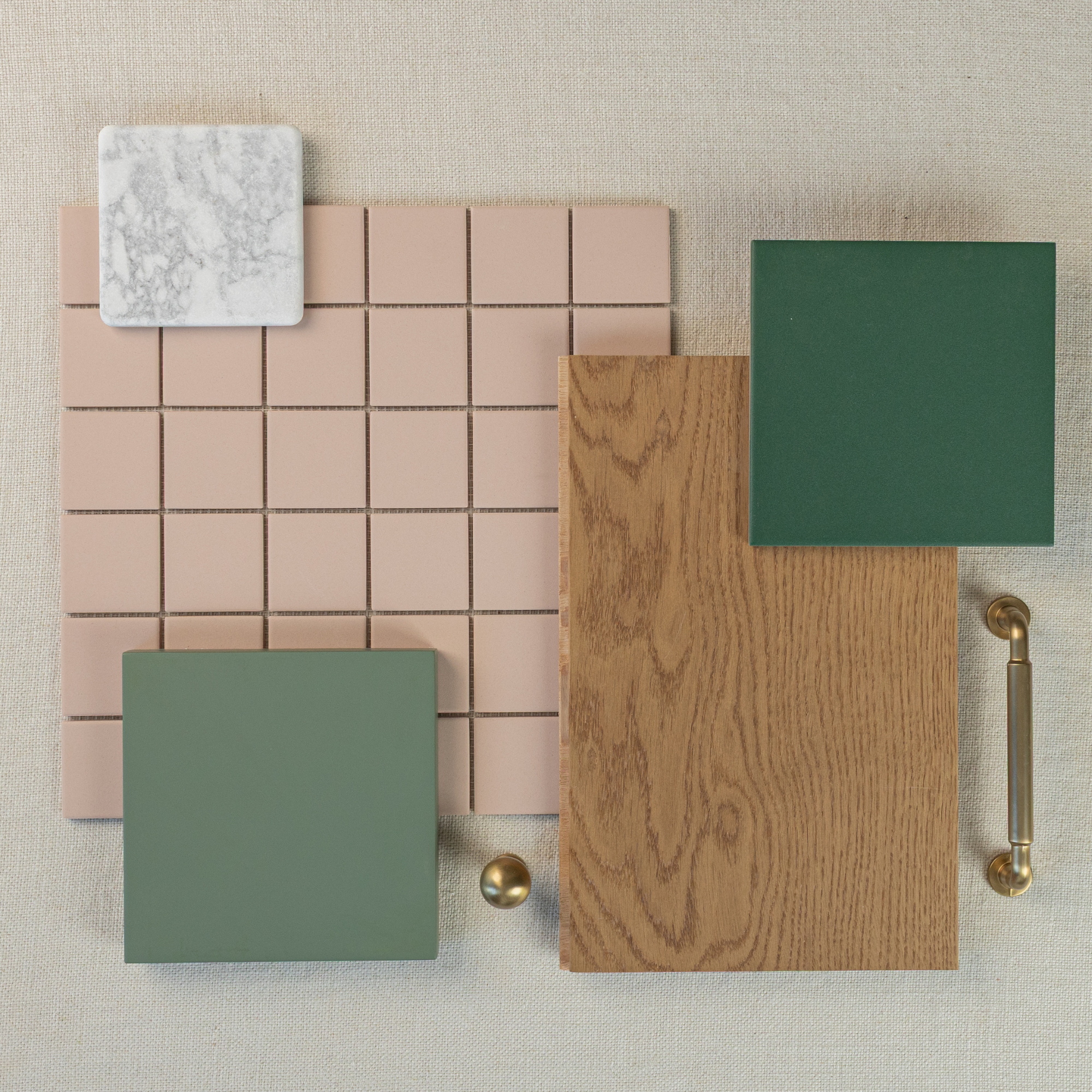 Flat lay of muted pink square mosaic tile with green ceramic tiles, wood sample, marble tile, brass knob, and handle on neutral fabric