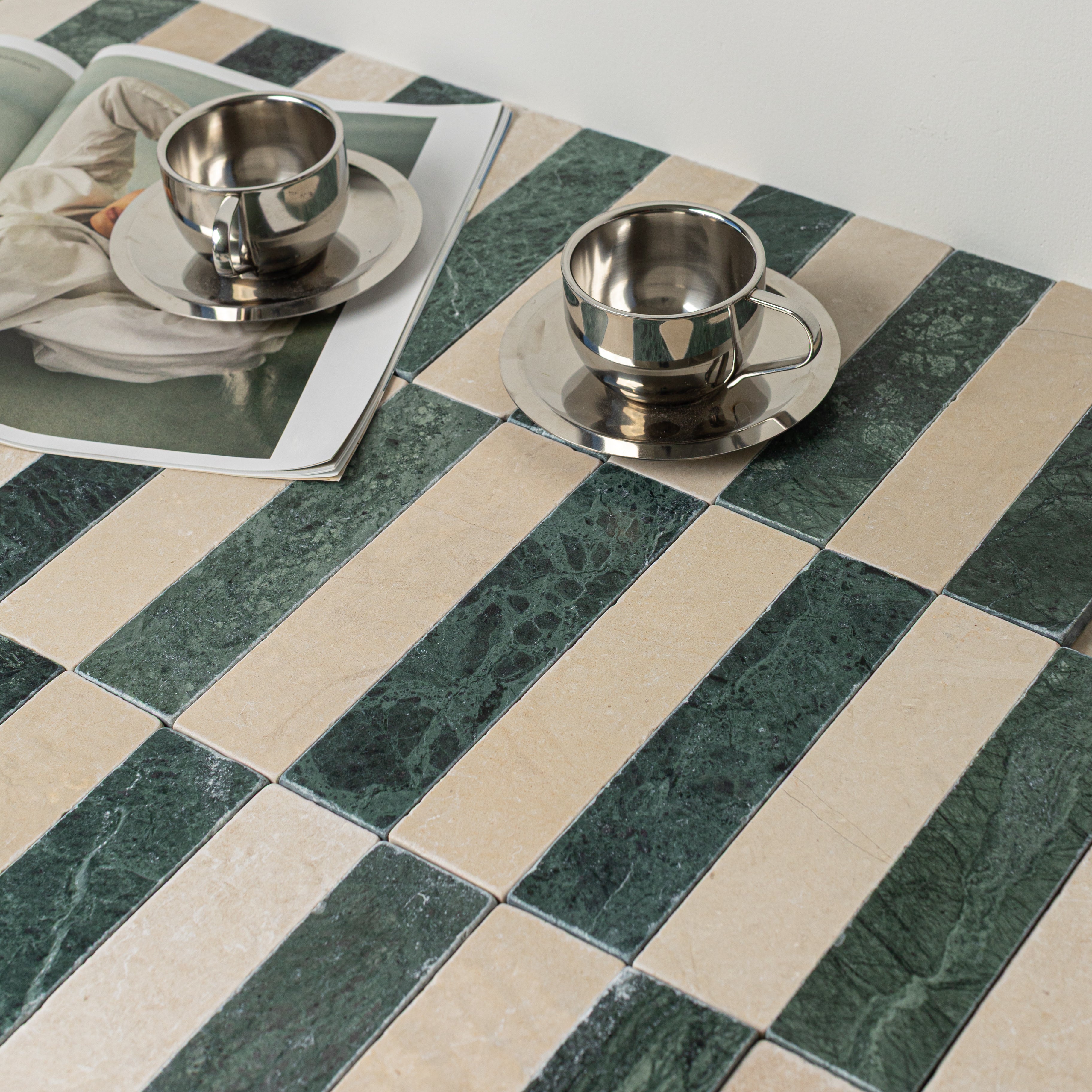 Green and beige rectangular porcelain tiles arranged in alternating stripes with two reflective metal cups on saucers and an open magazine nearby