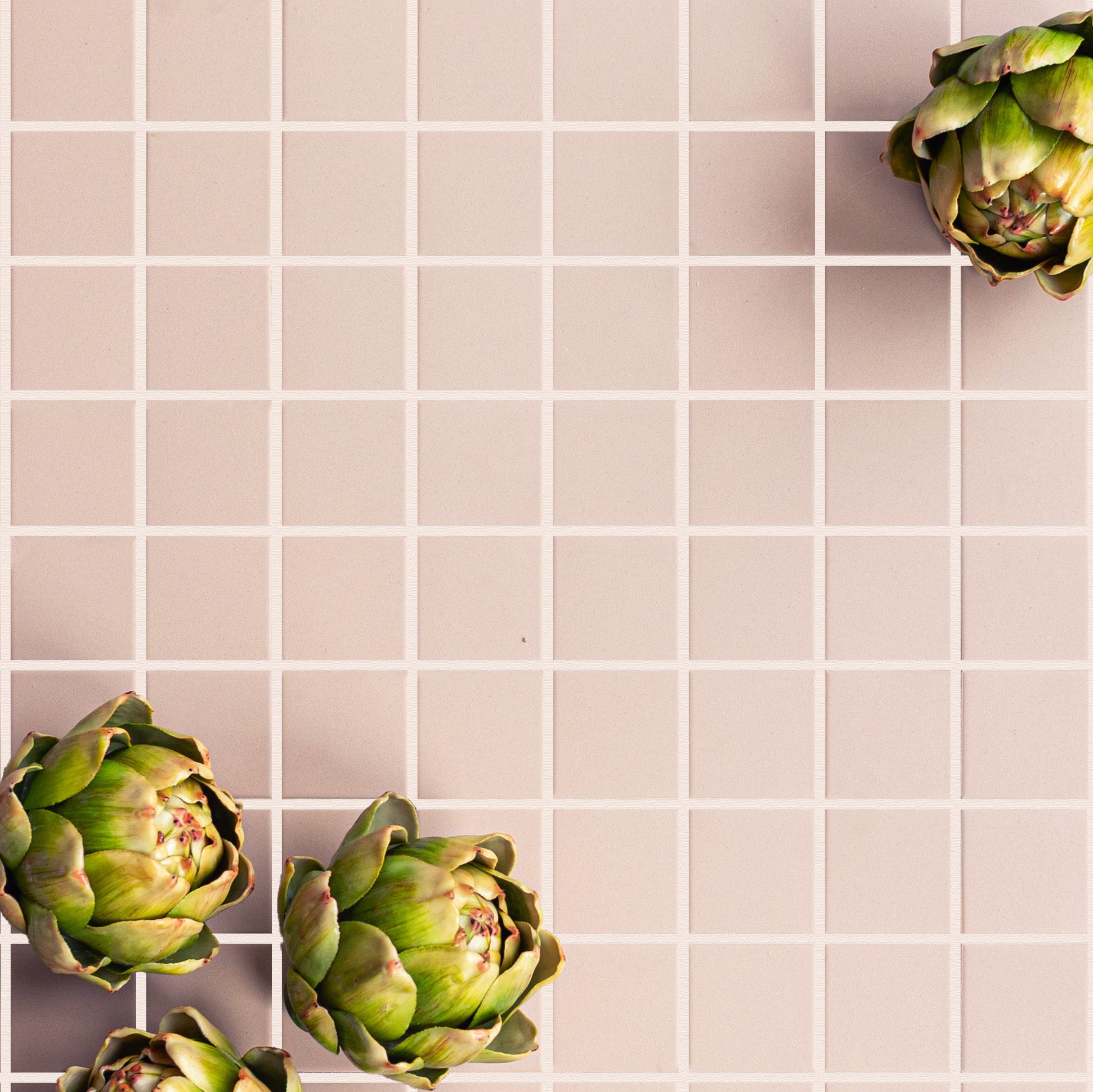 Soft beige small square ceramic tiles with clean grout lines and three fresh green artichokes placed on the surface