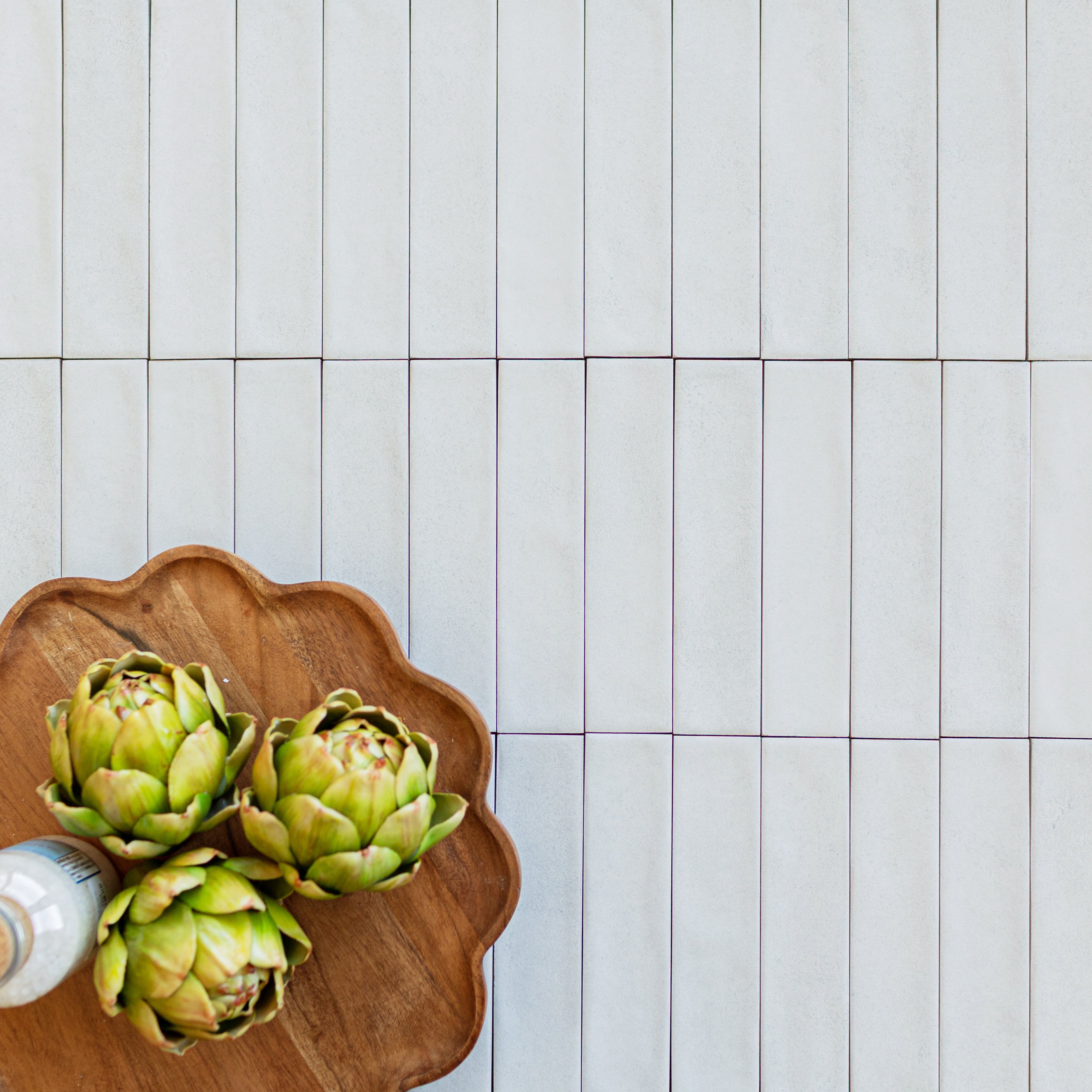 Soft white narrow ceramic tiles arranged vertically with a wooden tray holding three green artichokes nearby
