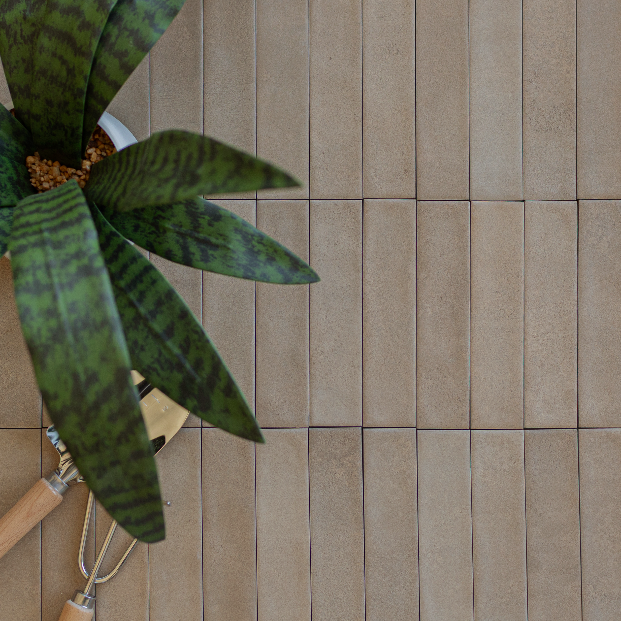 Long, narrow matte beige ceramic tiles arranged in staggered rows next to a potted green plant and gardening tools