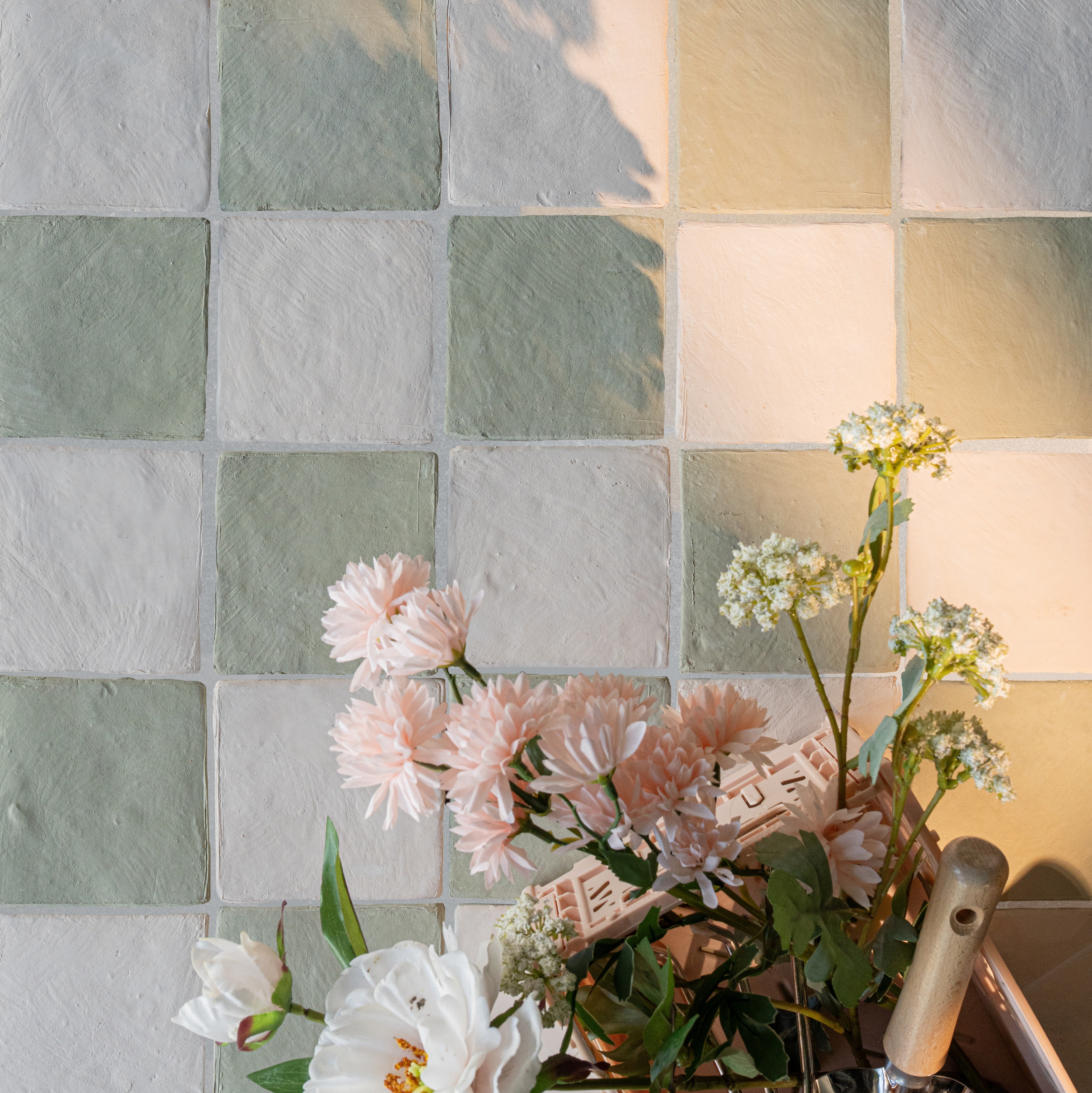 Soft green and cream ceramic tiles arranged in a checkered pattern beside delicate pink and white flowers in sunlight