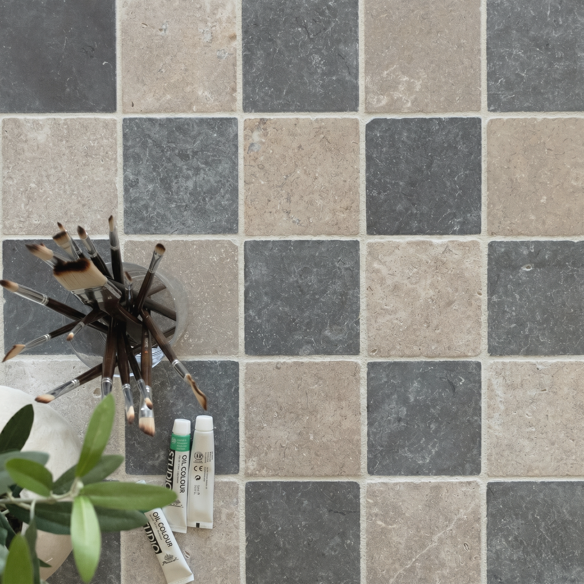 Checkerboard ceramic tile surface in muted grey and beige tones with paintbrushes and small paint tubes nearby