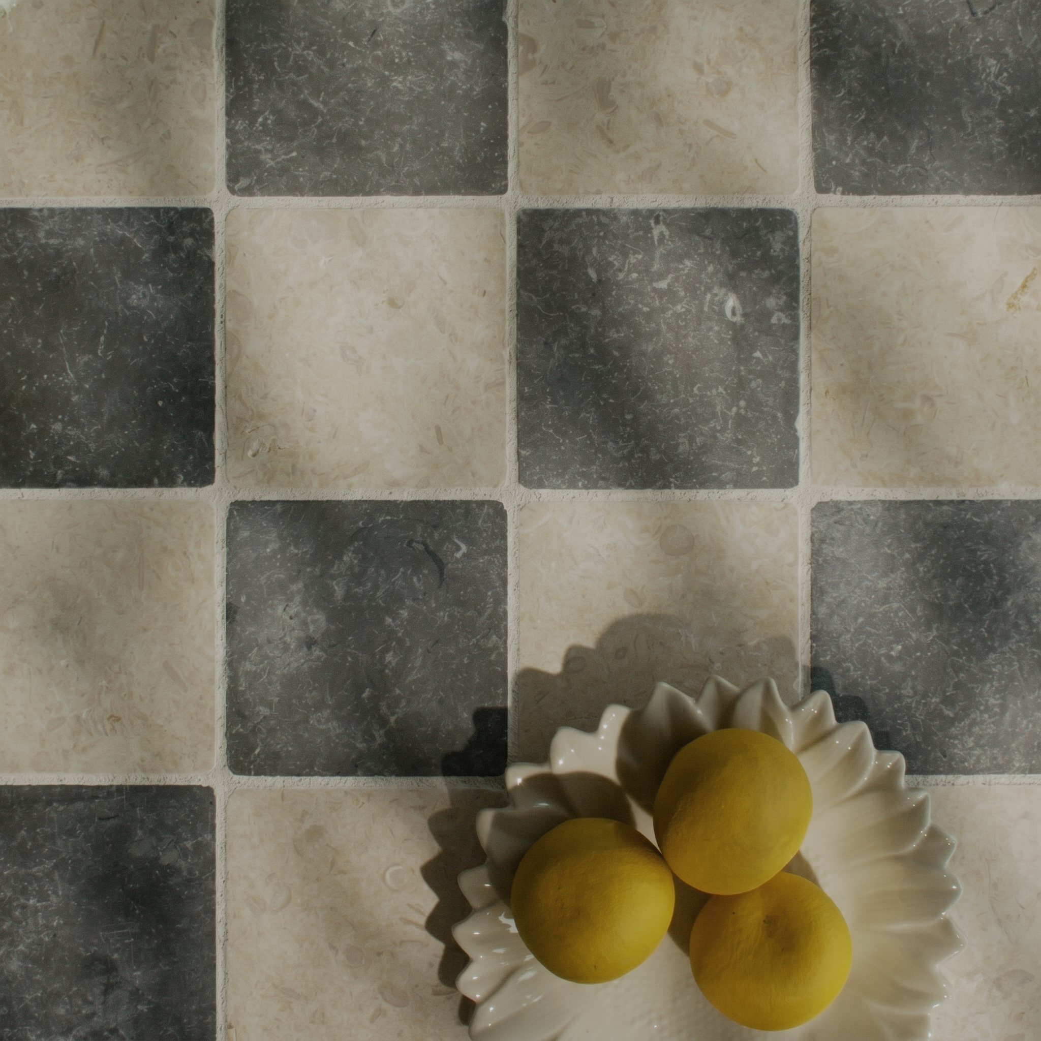 Checkerboard pattern of textured square tiles in soft beige and dark grey with a ceramic dish holding three yellow lemons.
