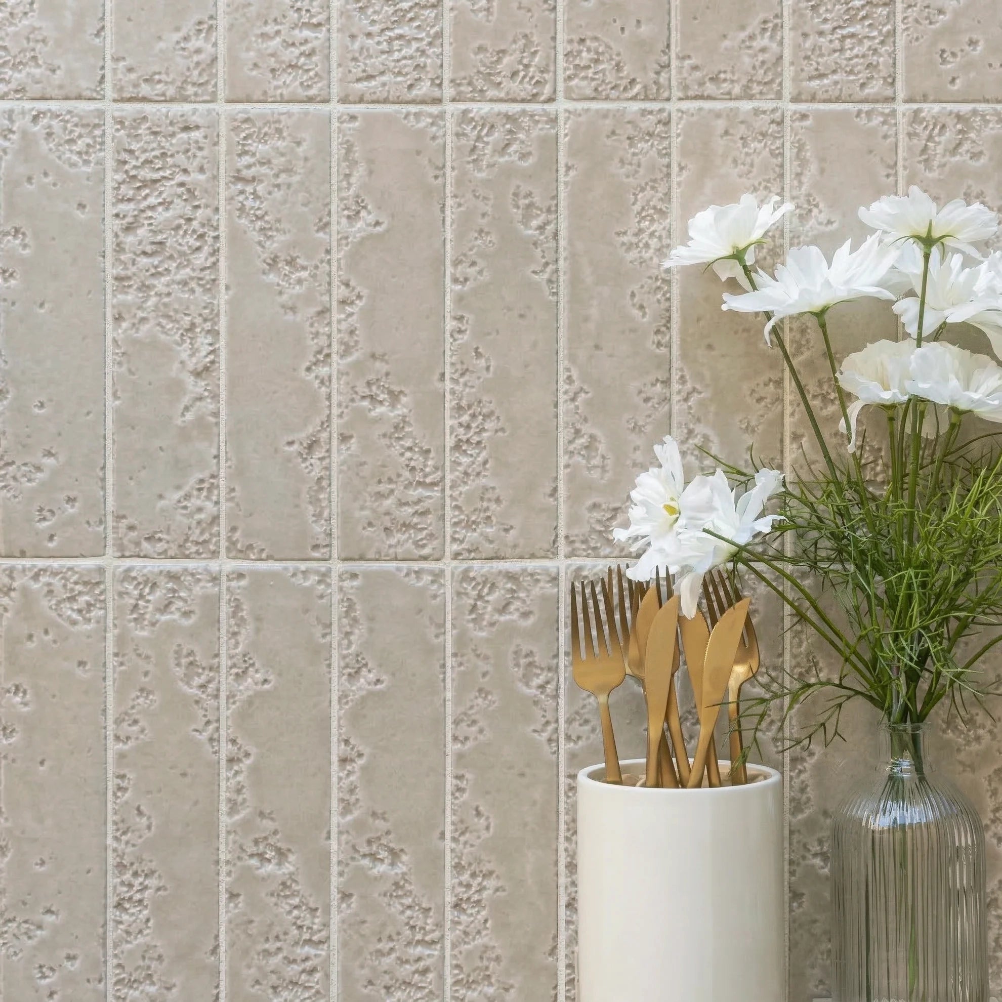 Vertical beige ceramic tiles with textured stone-effect surface next to a vase of white flowers and gold cutlery
