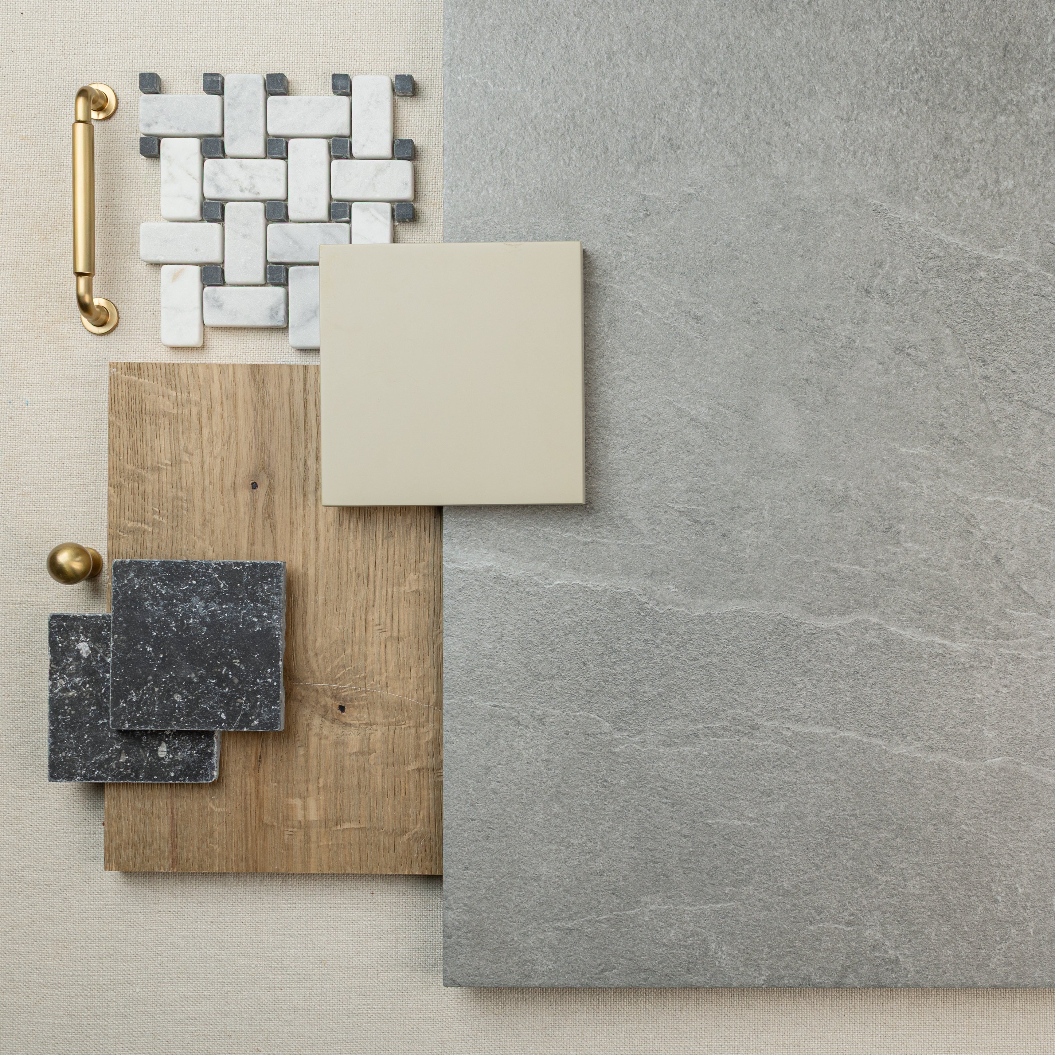 Flat lay of grey stone-look porcelain tile, wood sample, black speckled tiles, cream square tile, white mosaic, and brass hardware elements