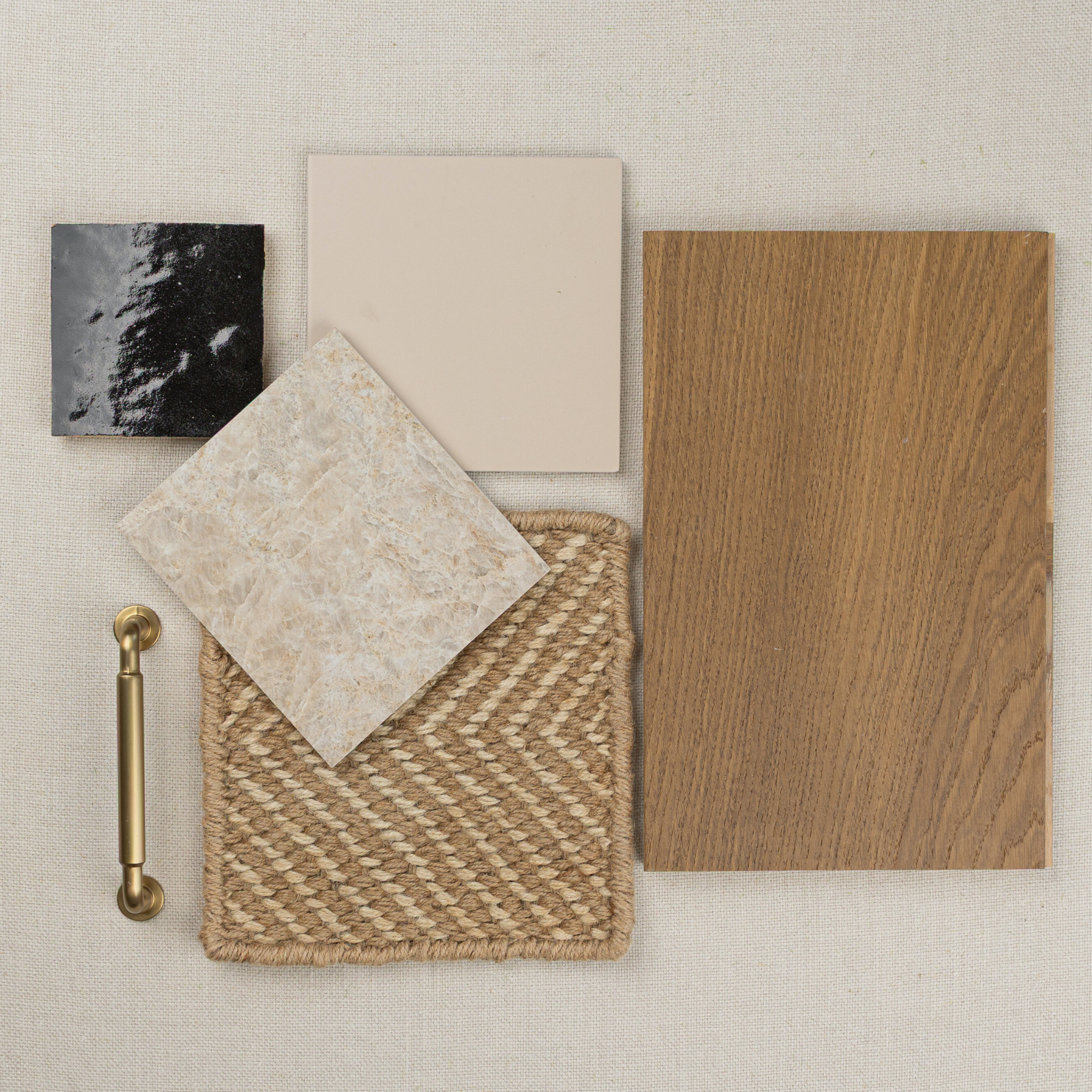 Top view of wood flooring sample, neutral and black ceramic tiles, woven placemat, and brass drawer pull on beige fabric background