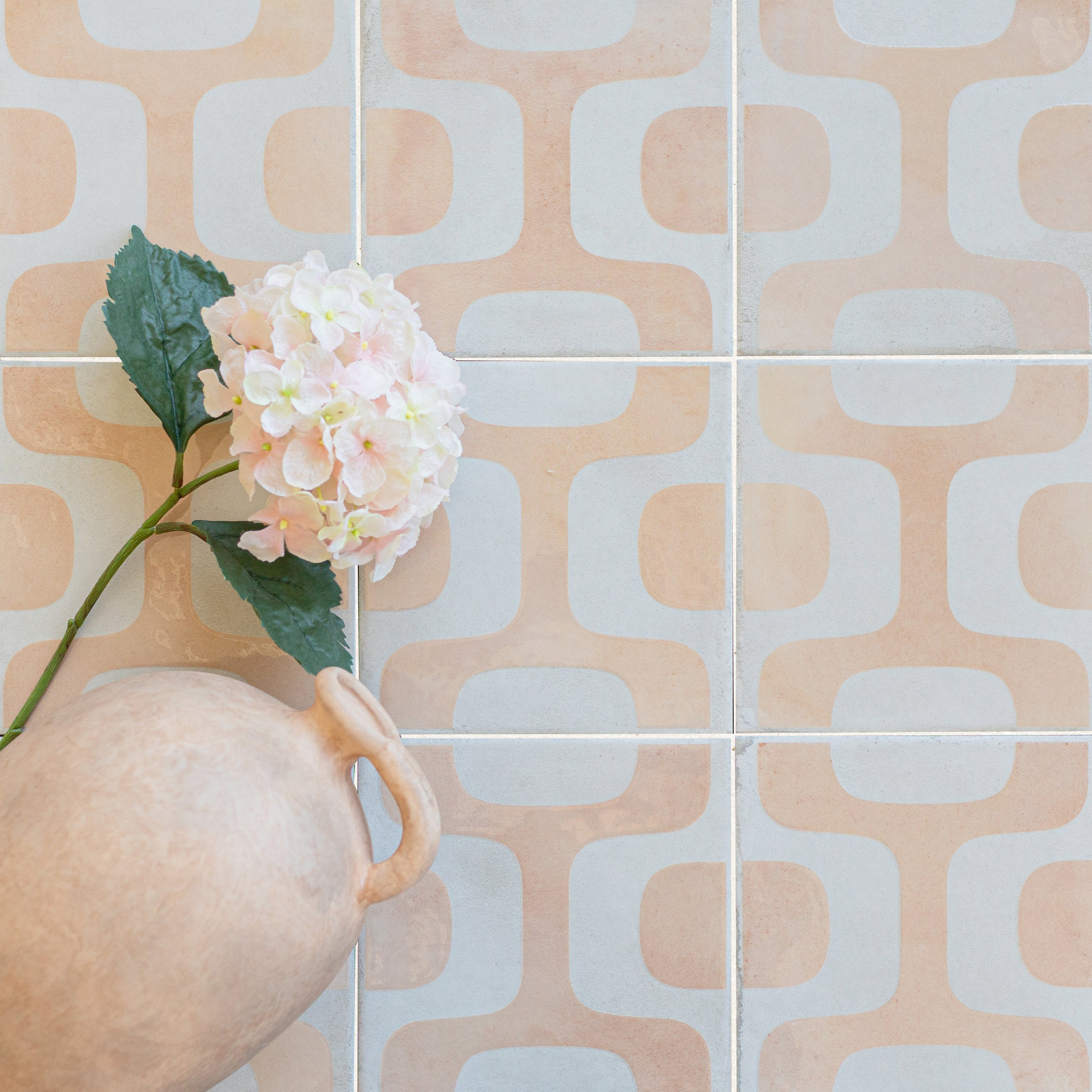 Square porcelain tiles in soft peach and light gray tones with retro curved patterns beside a clay pitcher and pink flower