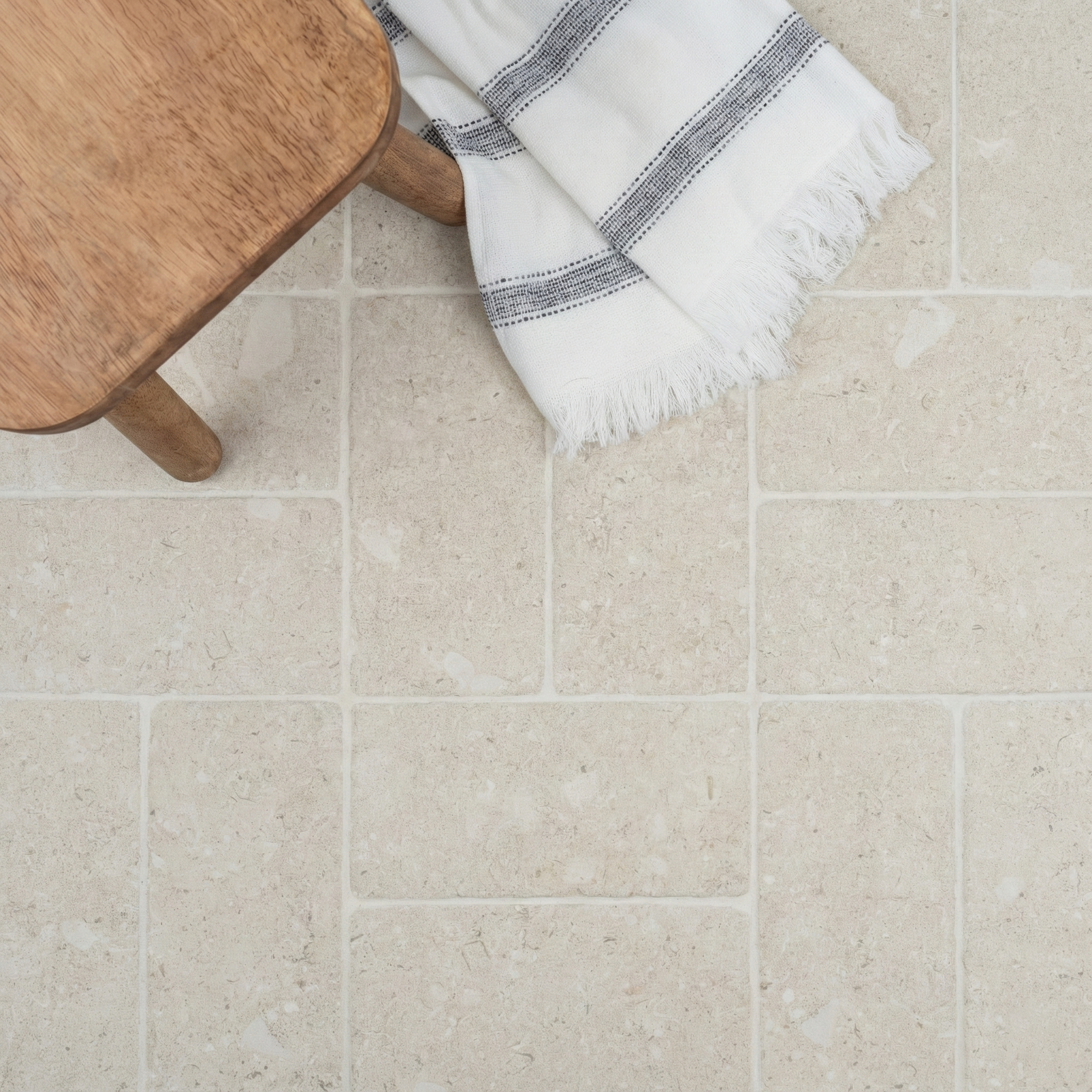 Overhead view of light ivory ceramic tiles with natural stone patterns next to a wooden stool and fringed striped cloth