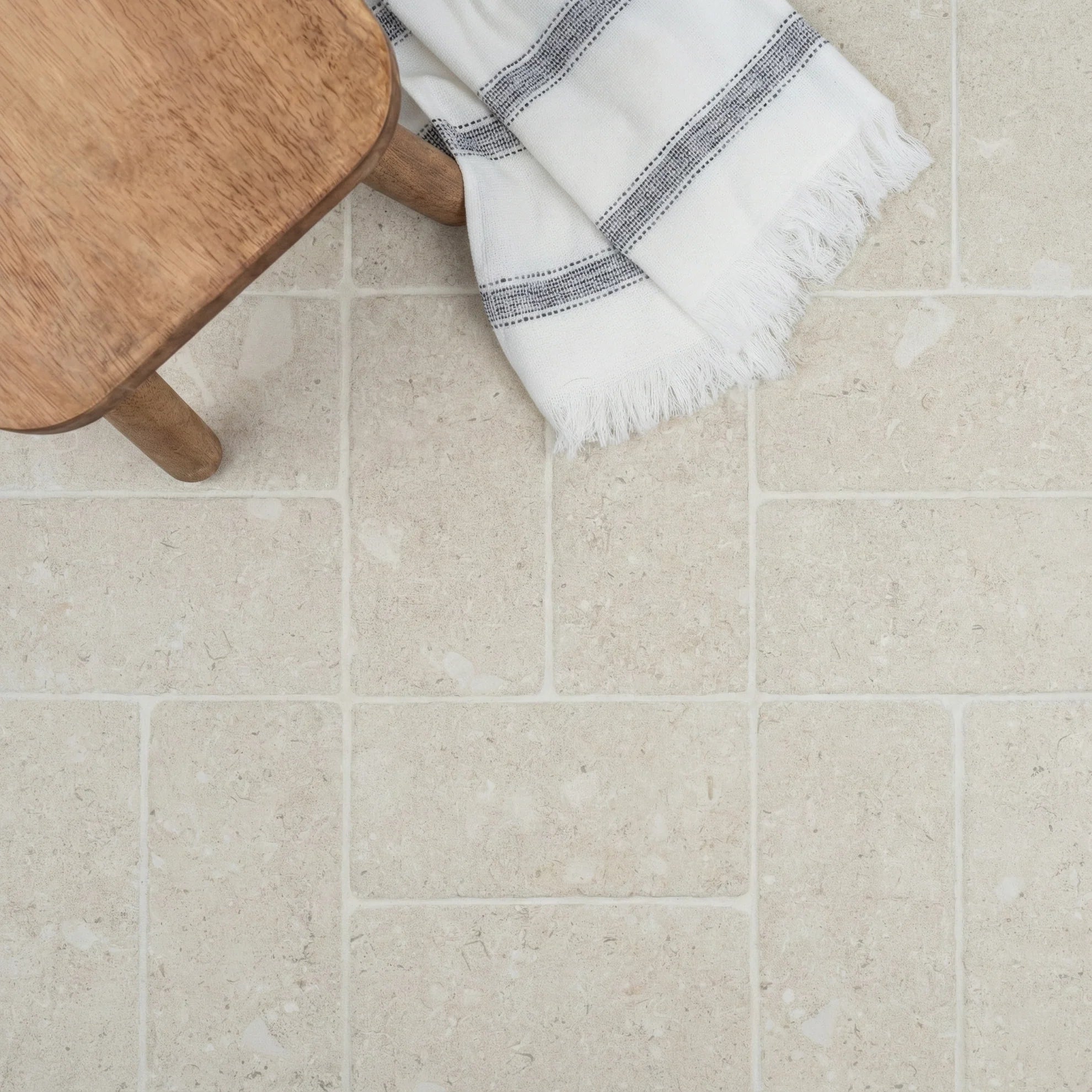 Matte ivory ceramic tiles with subtle texture arranged with a wooden stool leg and fringed white striped fabric nearby