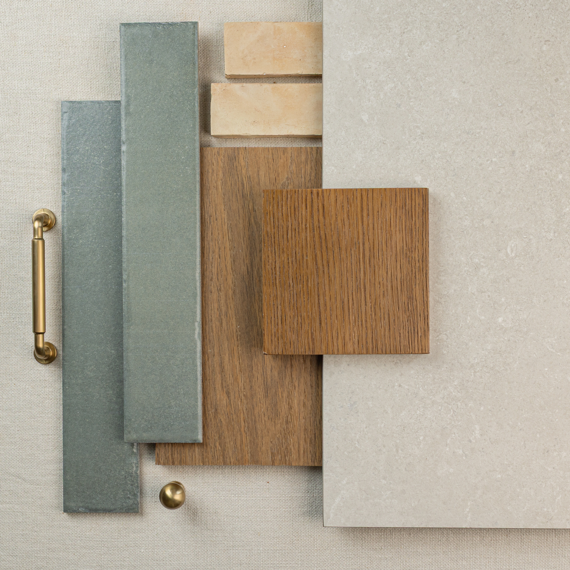 Flat lay of green ceramic tiles, light wood plank, beige stone tiles, and brass hardware on textured fabric background