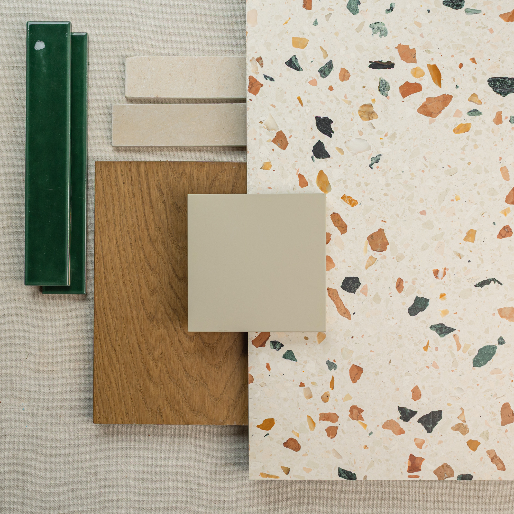Flat lay of warm wood, beige tiles, speckled terrazzo, and glossy deep green ceramic samples on neutral fabric background
