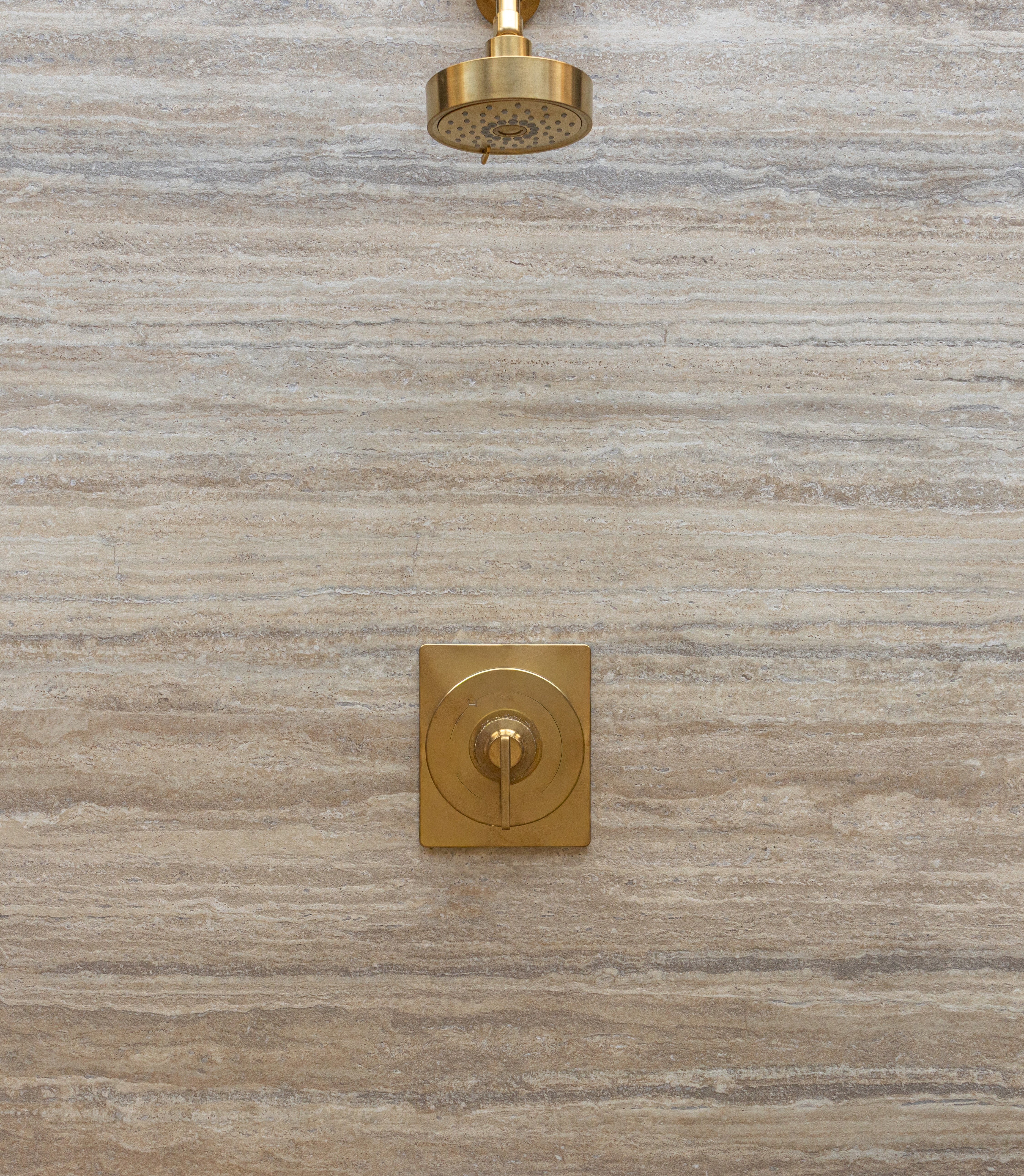 Brushed gold showerhead and control mounted on a large natural stone-effect tile with horizontal striations