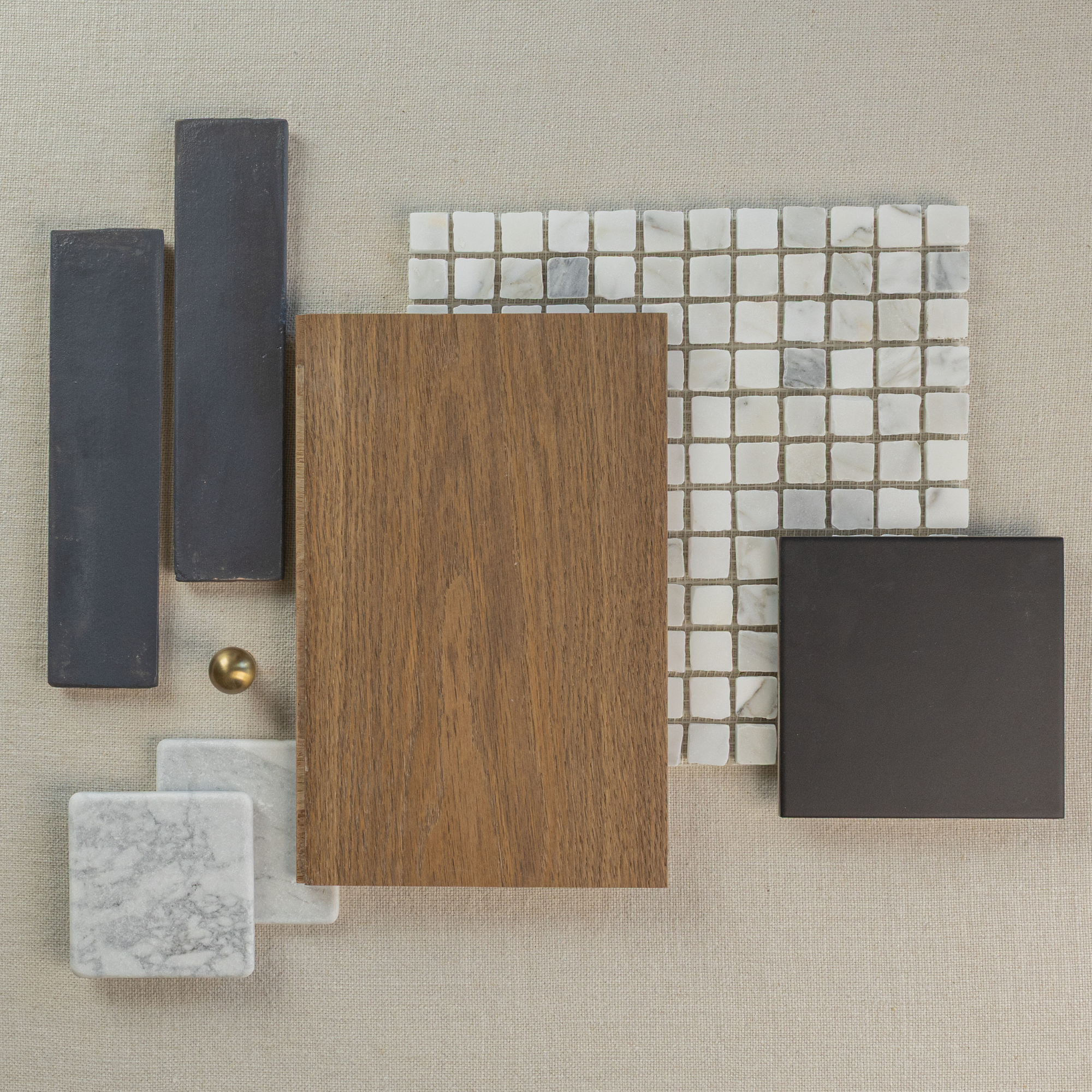 Flat lay of wood plank, white marble mosaic tiles, dark rectangular and square tiles, and small brass hardware on beige fabric