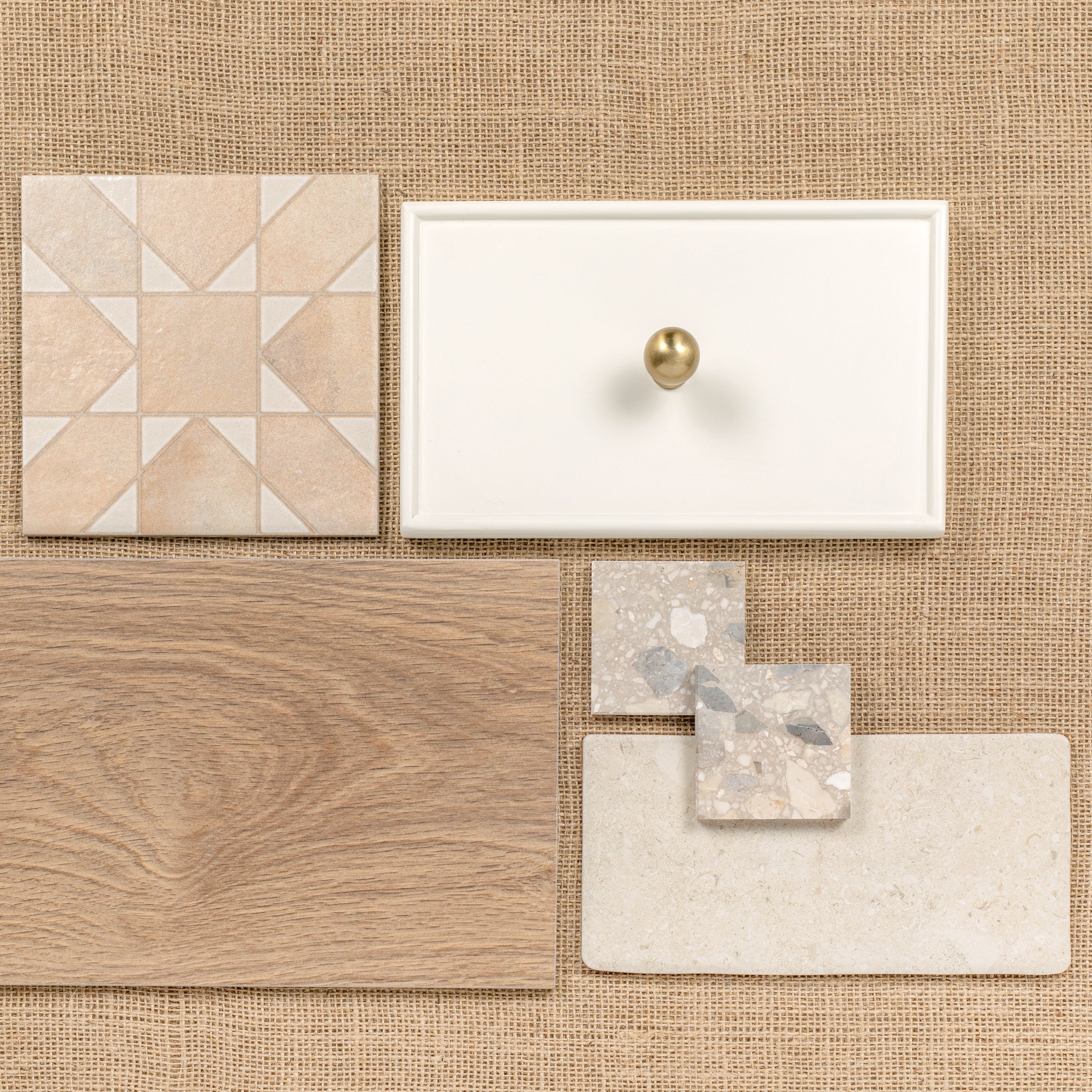 Flat lay of ceramic tile, wood plank, terrazzo samples, and white cabinet drawer front with brass knob on burlap surface