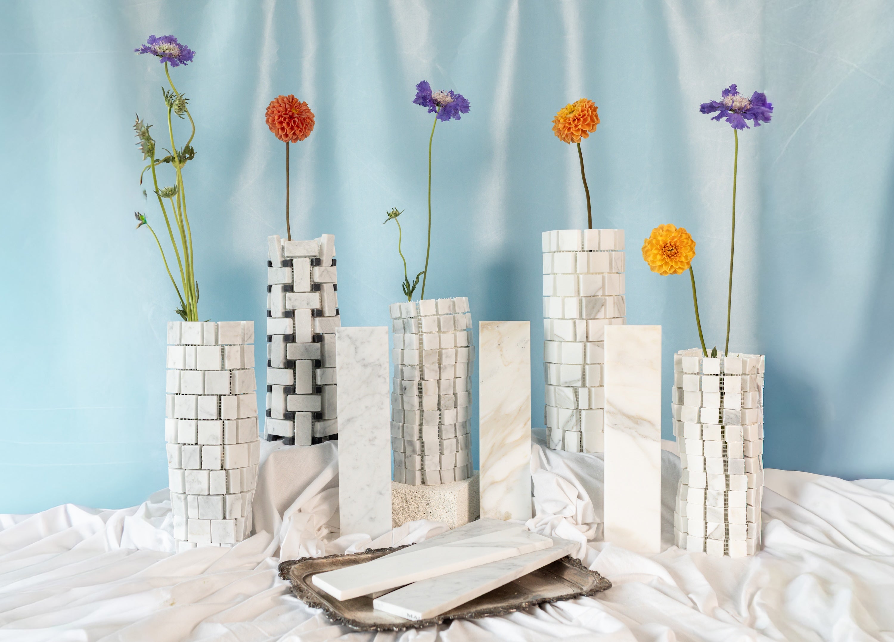 Tall cylindrical white marble vases with geometric tile patterns holding colorful flowers against a blue backdrop