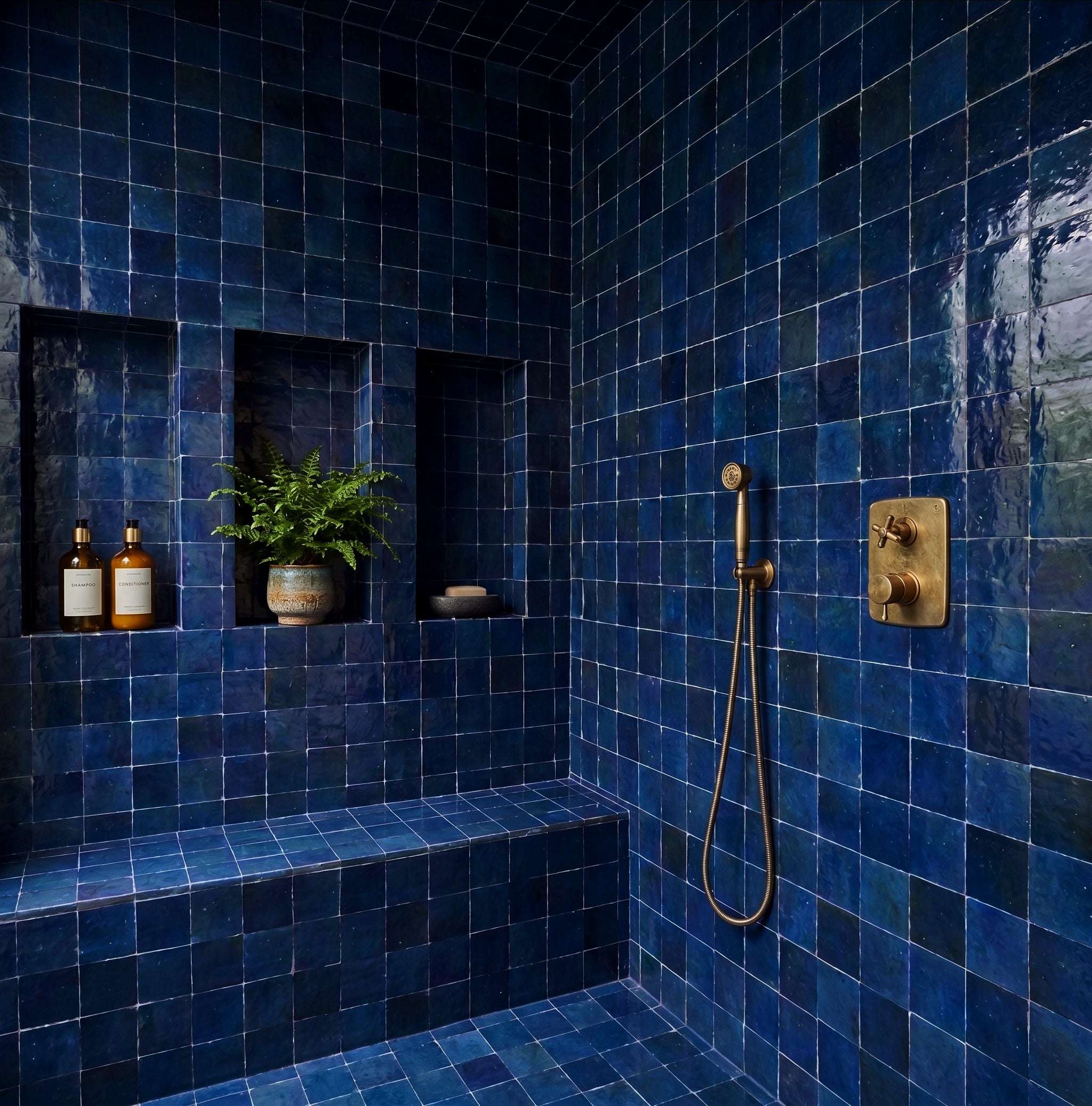 Bathroom shower with deep blue handmade ceramic small format Midnight Glazed Zellige Tile and brass fixtures, niche with plants and bottles.
