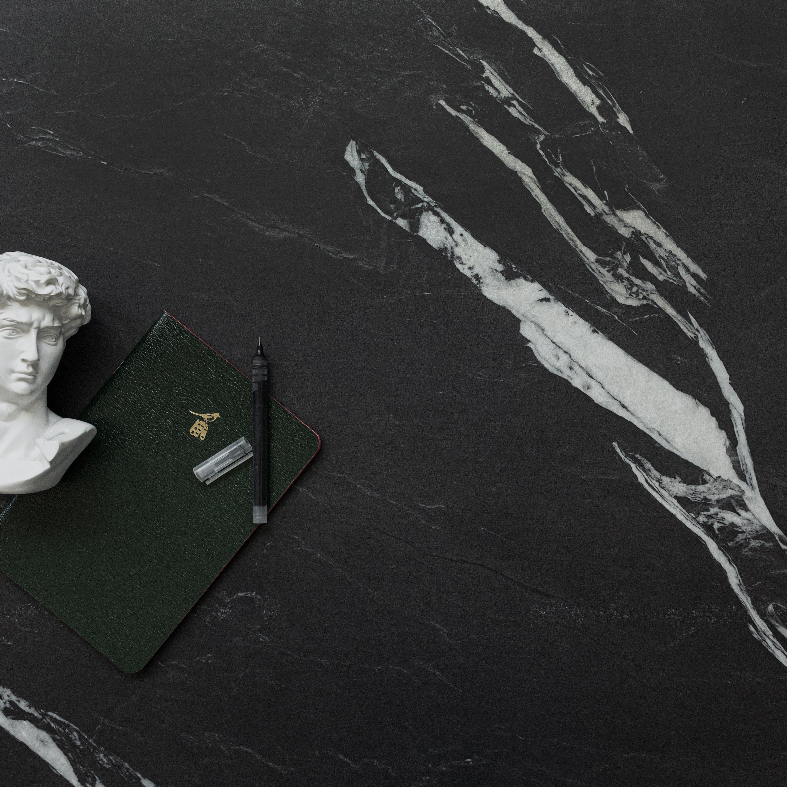 Dark porcelain tile with bold white marble-like veining, styled with a small classical bust, notebook, and pen.