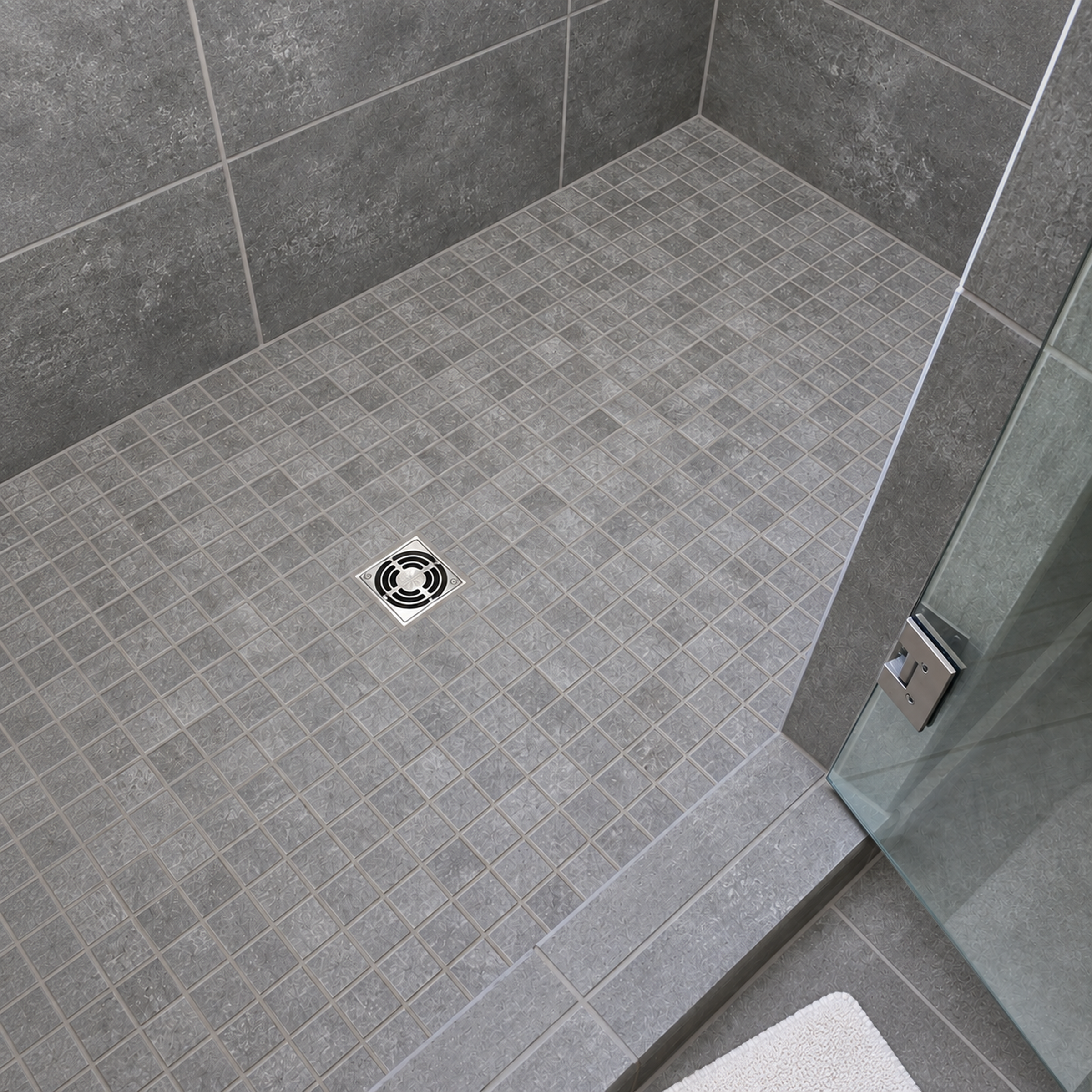 Gray tiled walk-in shower with mosaic floor square drain glass door and white bath mat
