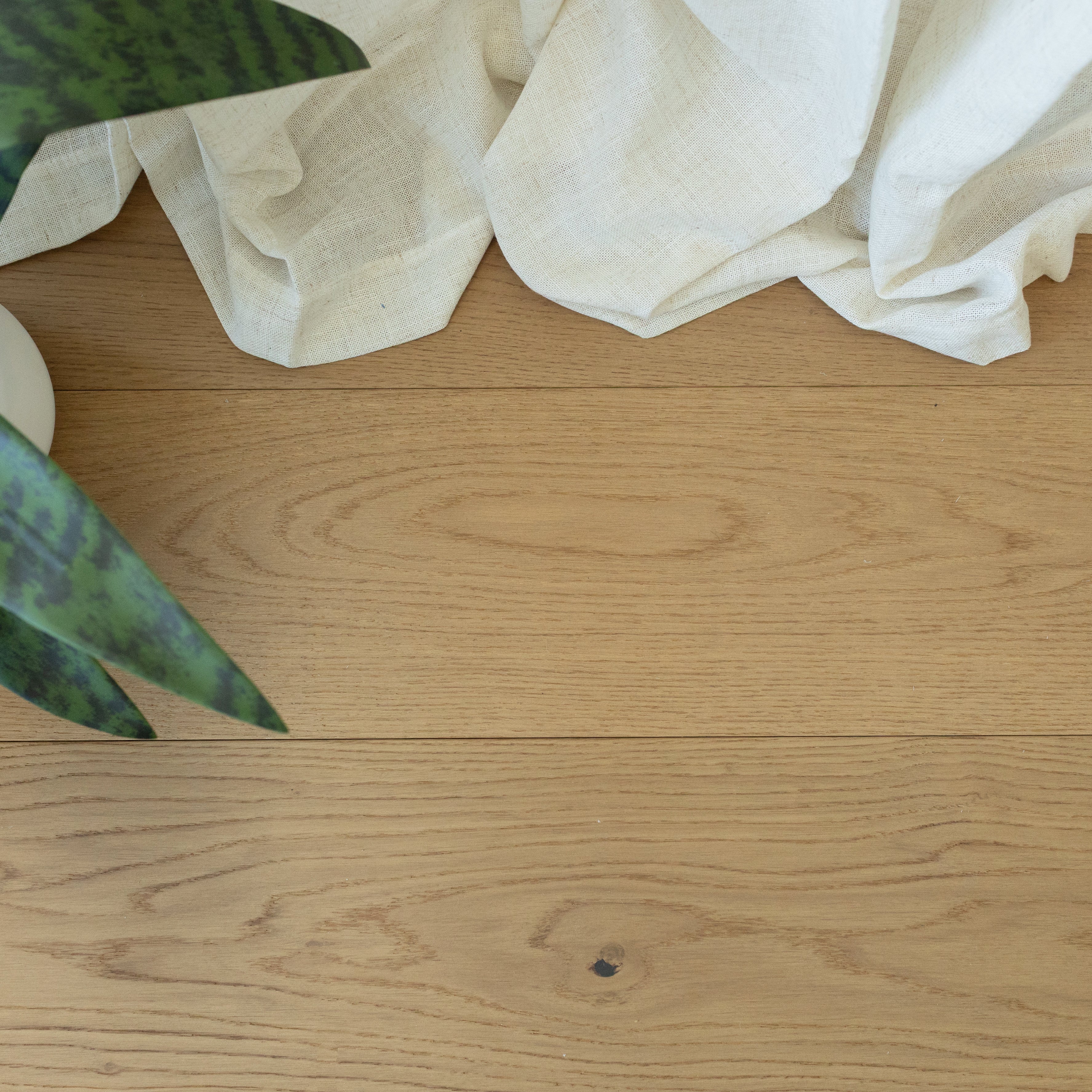 Natural oak engineered wood flooring with soft beige linen fabric and a green plant leaf peeking in the corner
