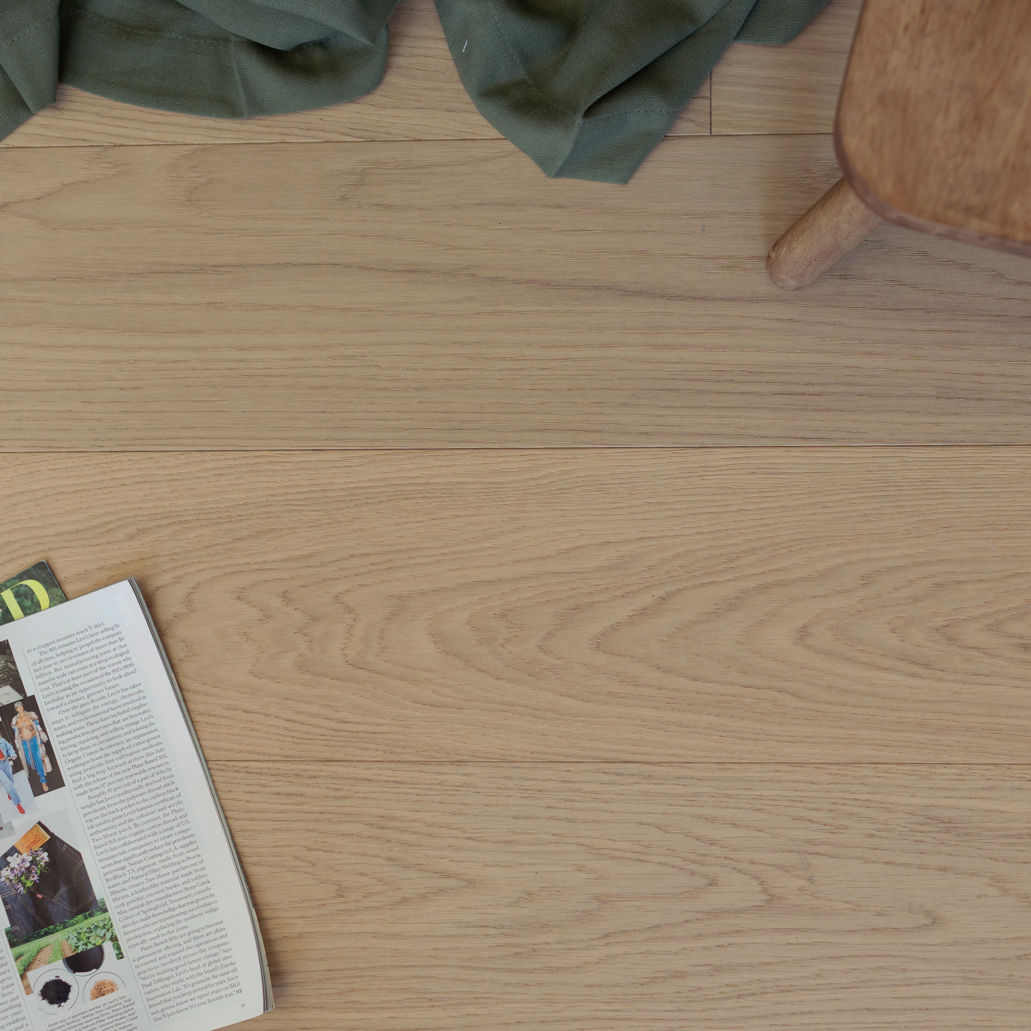 Natural oak wood flooring with visible grain texture, a corner of a wooden chair, green fabric, and an open magazine on floor
