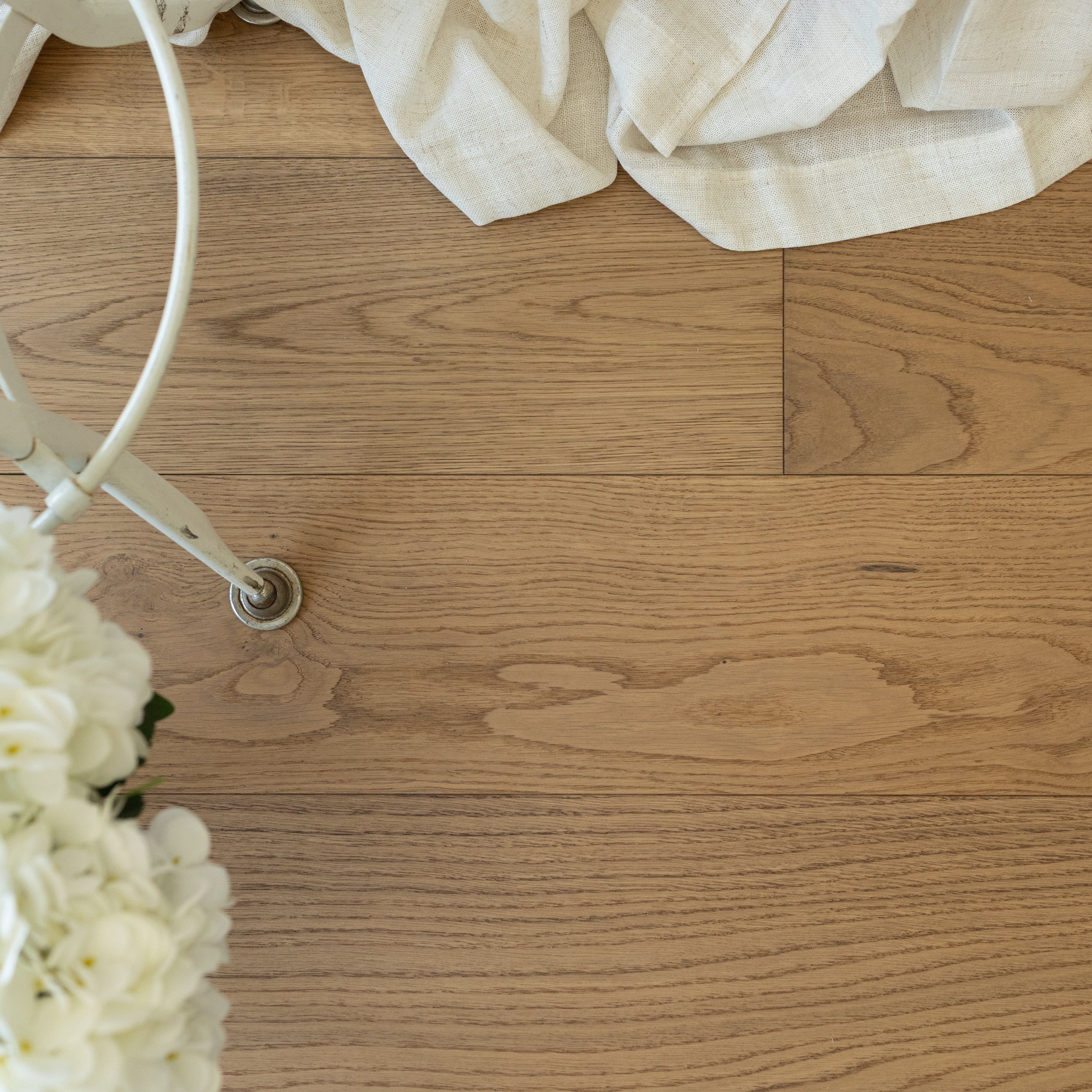 Light warm oak wood flooring with natural grain under soft beige fabric and white hydrangea flowers near metal chair leg