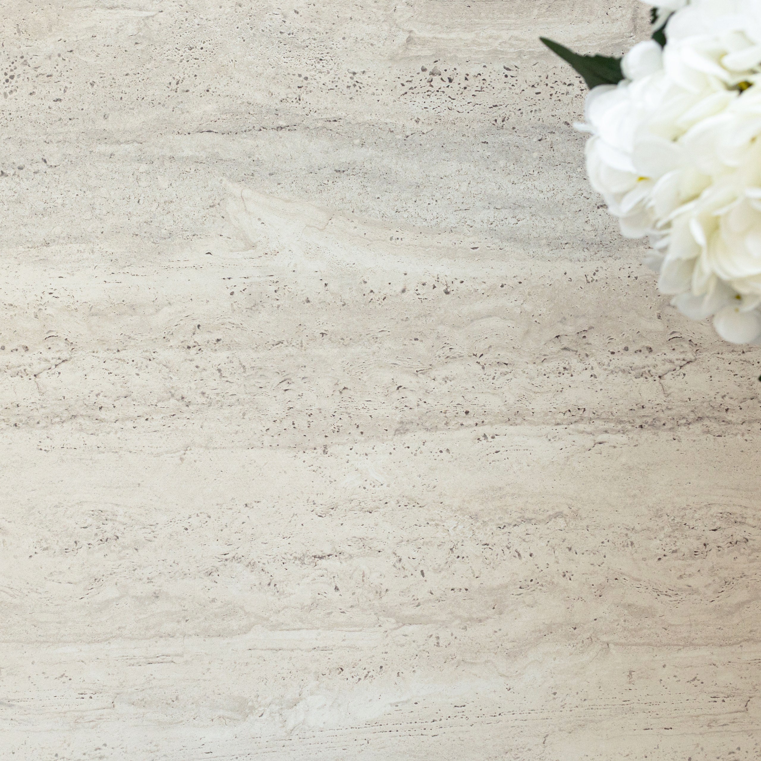 Close-up of light beige travertine porcelain tile with natural horizontal veining and white flowers in the corner