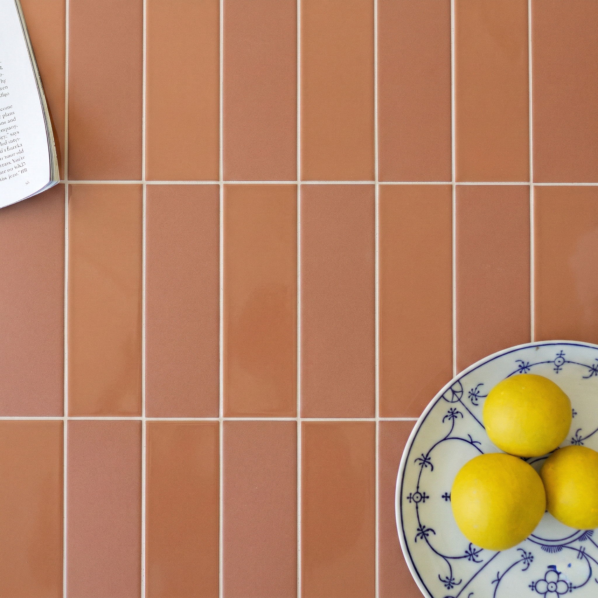 Vertical rectangular orange porcelain tiles with white grout next to a plate holding three lemons and an open book page