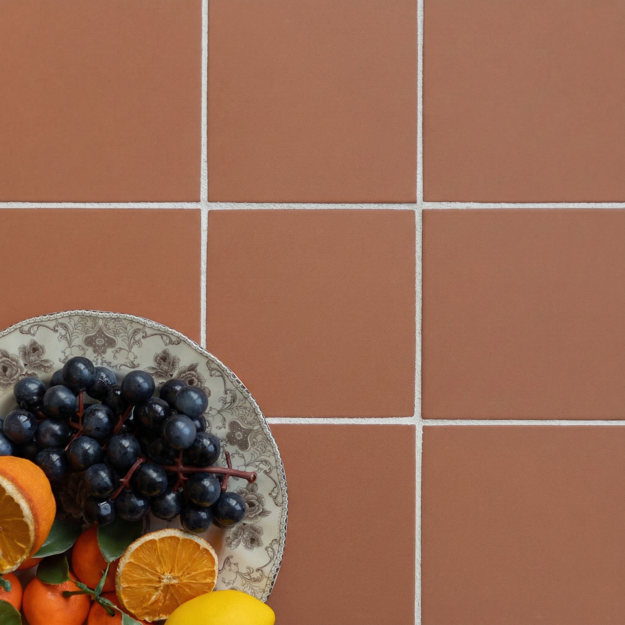 Orange tiles with white grout surrounding a plate of grapes, citrus, and persimmons on a tile floor surface