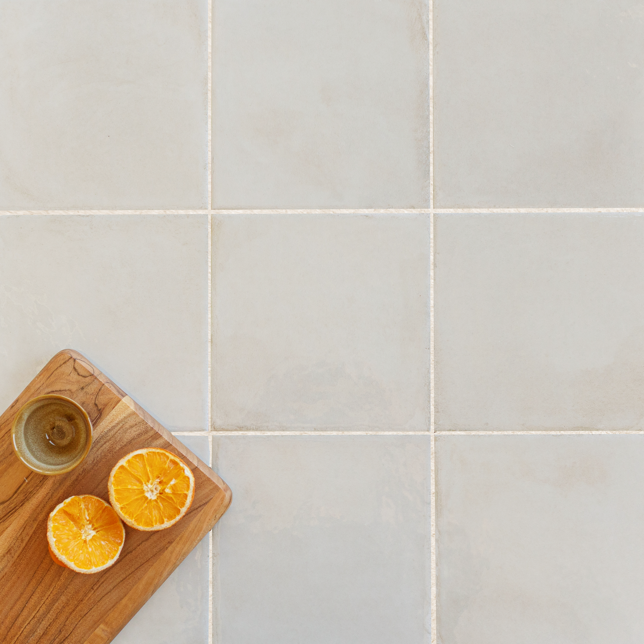 Muted beige ceramic tiles arranged in a grid with a wooden board holding two orange halves and a small bowl nearby