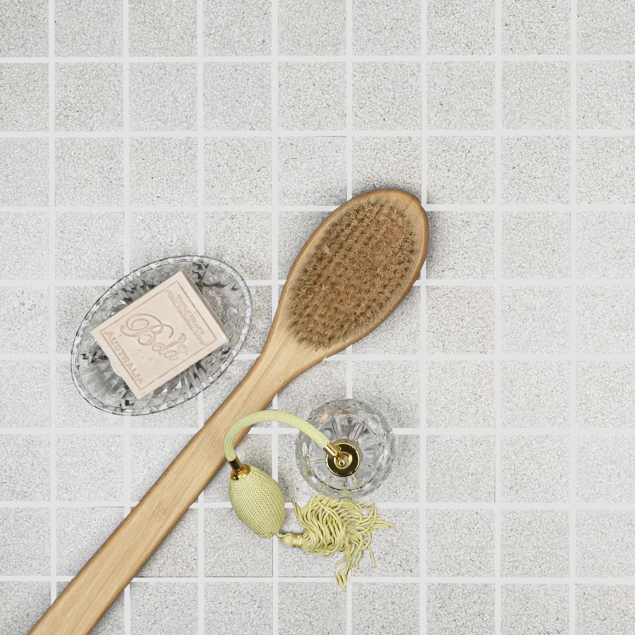 Grid of small light gray porcelain tiles with subtle texture beneath a wooden bath brush, vintage perfume atomizer, and soap dish