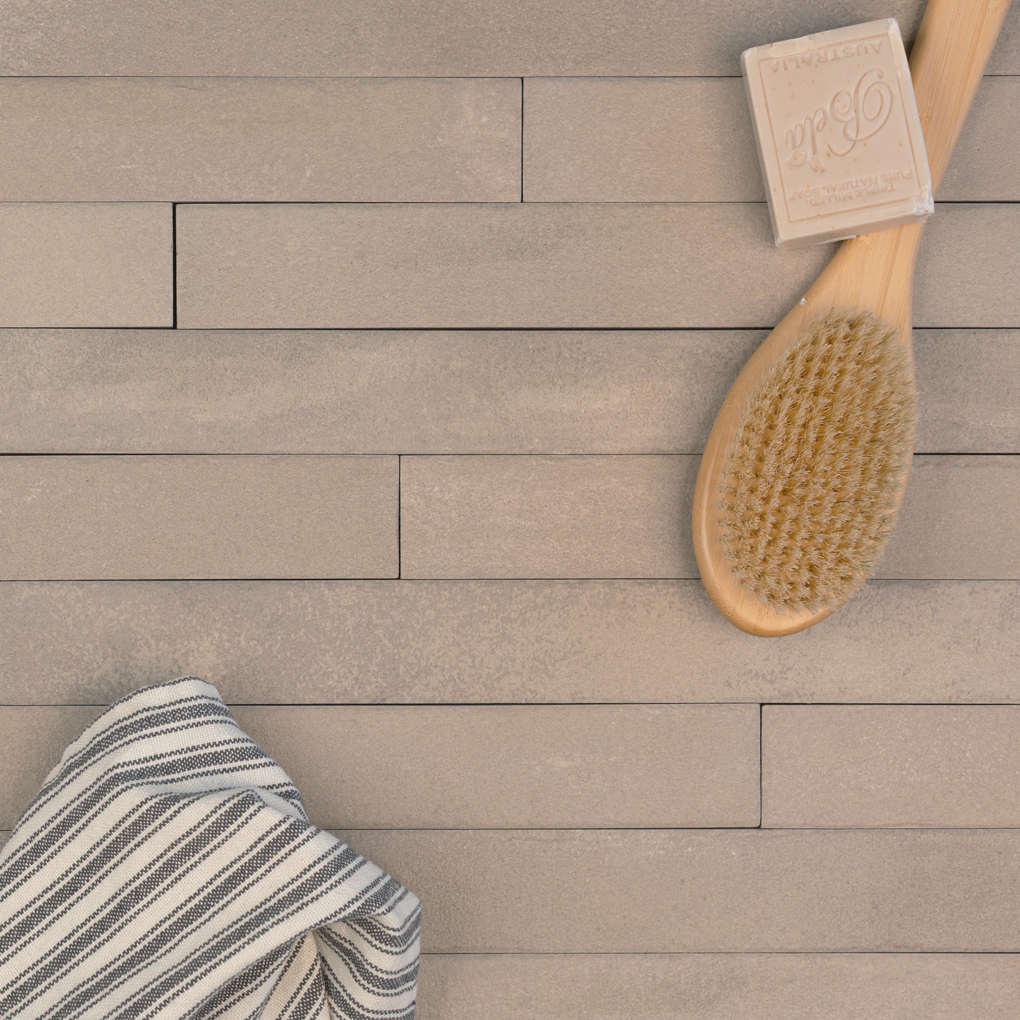 Matte beige rectangular ceramic tiles arranged in staggered rows with a wooden brush, soap bar, and striped cloth nearby