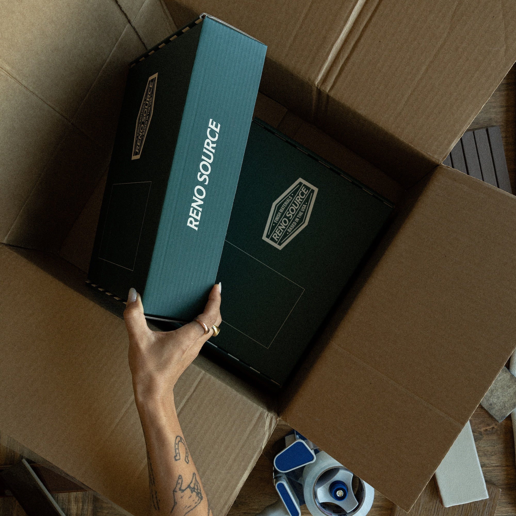 Hand reaching into a cardboard box to lift a dark green Reno Source sample folder amid packing materials on a wooden floor