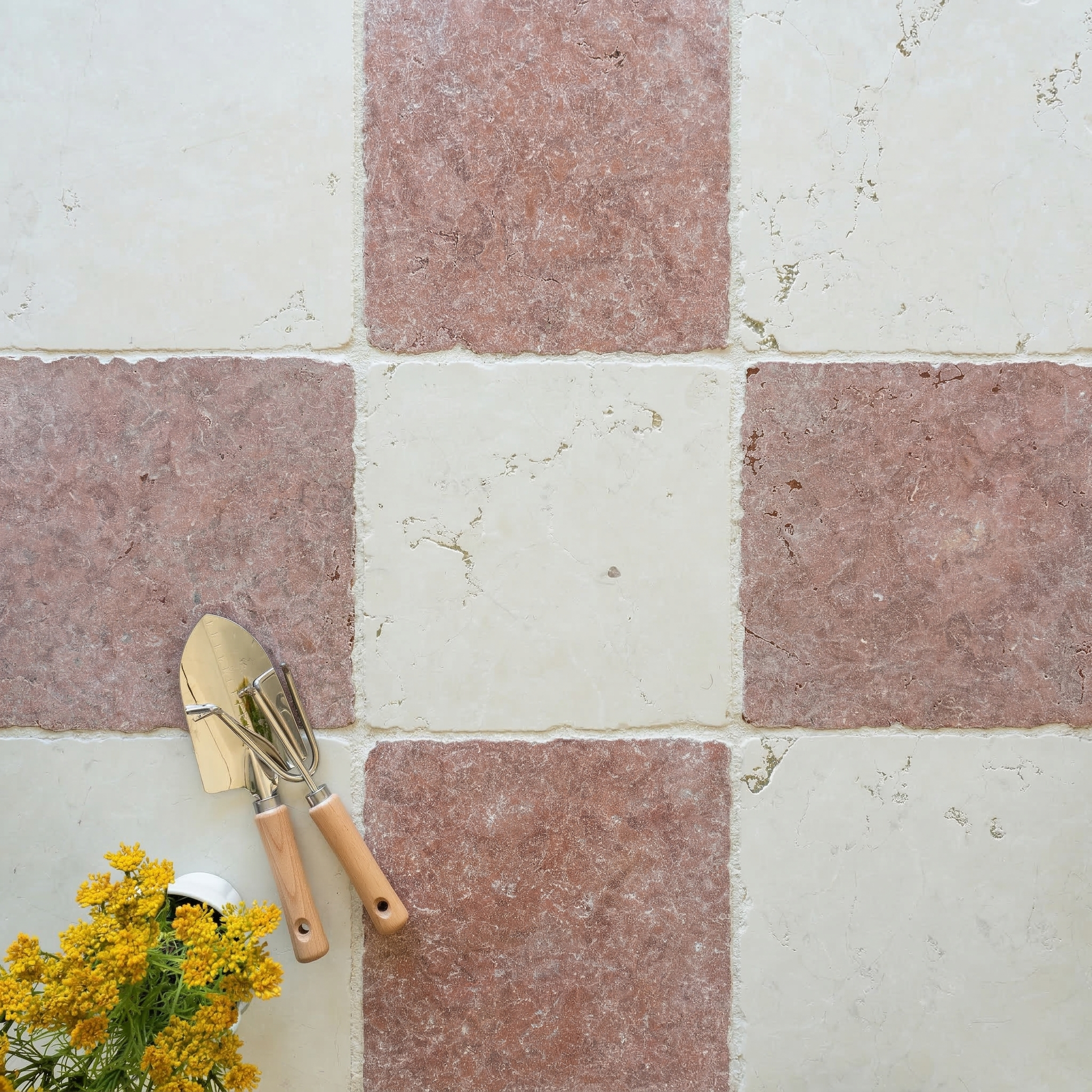 Checkerboard pattern of tumbled stone tiles in soft red and cream tones with garden tools and yellow flowers nearby