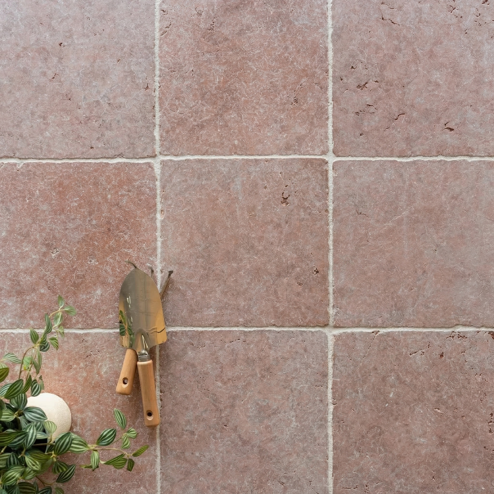 Square terracotta tiles with subtle texture arranged in a grid beside small garden trowels and green foliage leaves