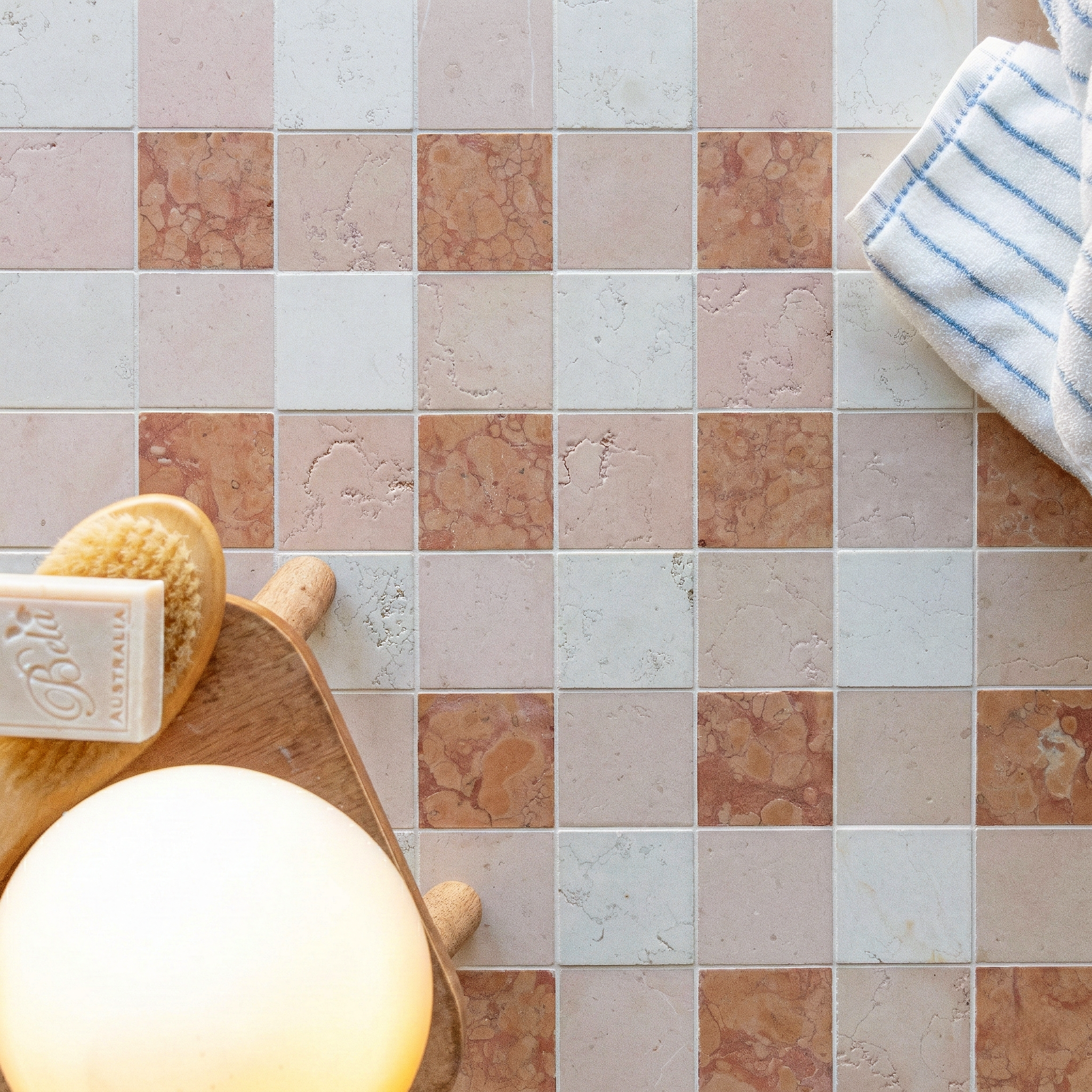 Checkerboard arrangement of pink, beige, and cream ceramic tiles beside wooden brush, soap, and striped cloth