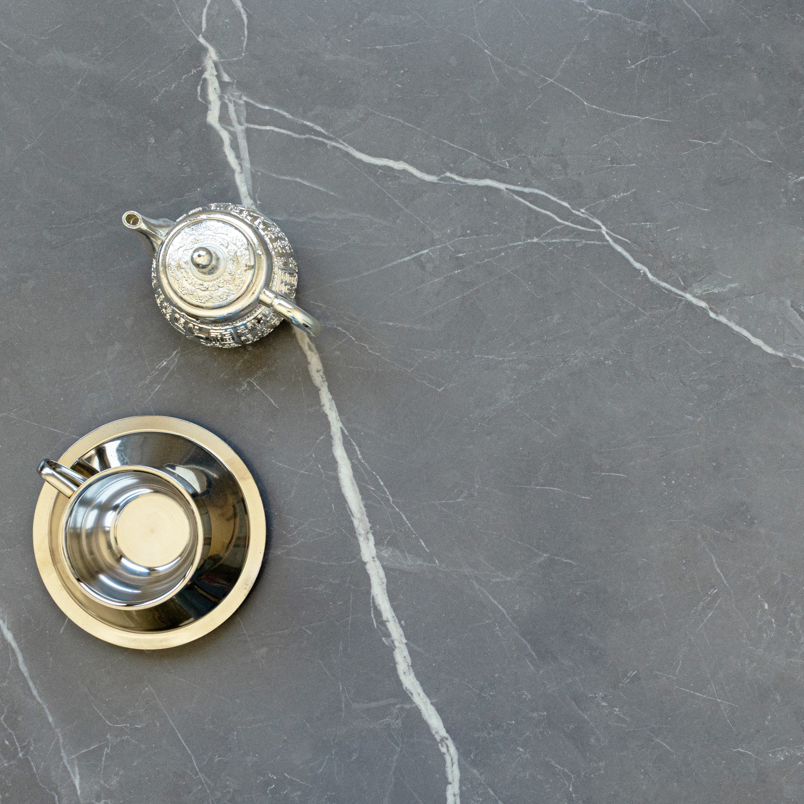 Matte gray porcelain surface with white marble-like veining featuring a silver teapot and cup on saucer placed above