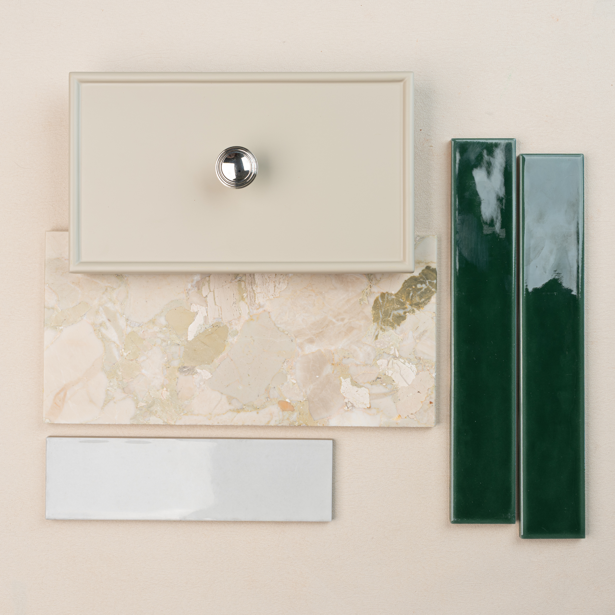 Flat lay of cream drawer front with silver knob, beige stone tile, glossy dark green ceramic tiles, and white rectangular tile on light background