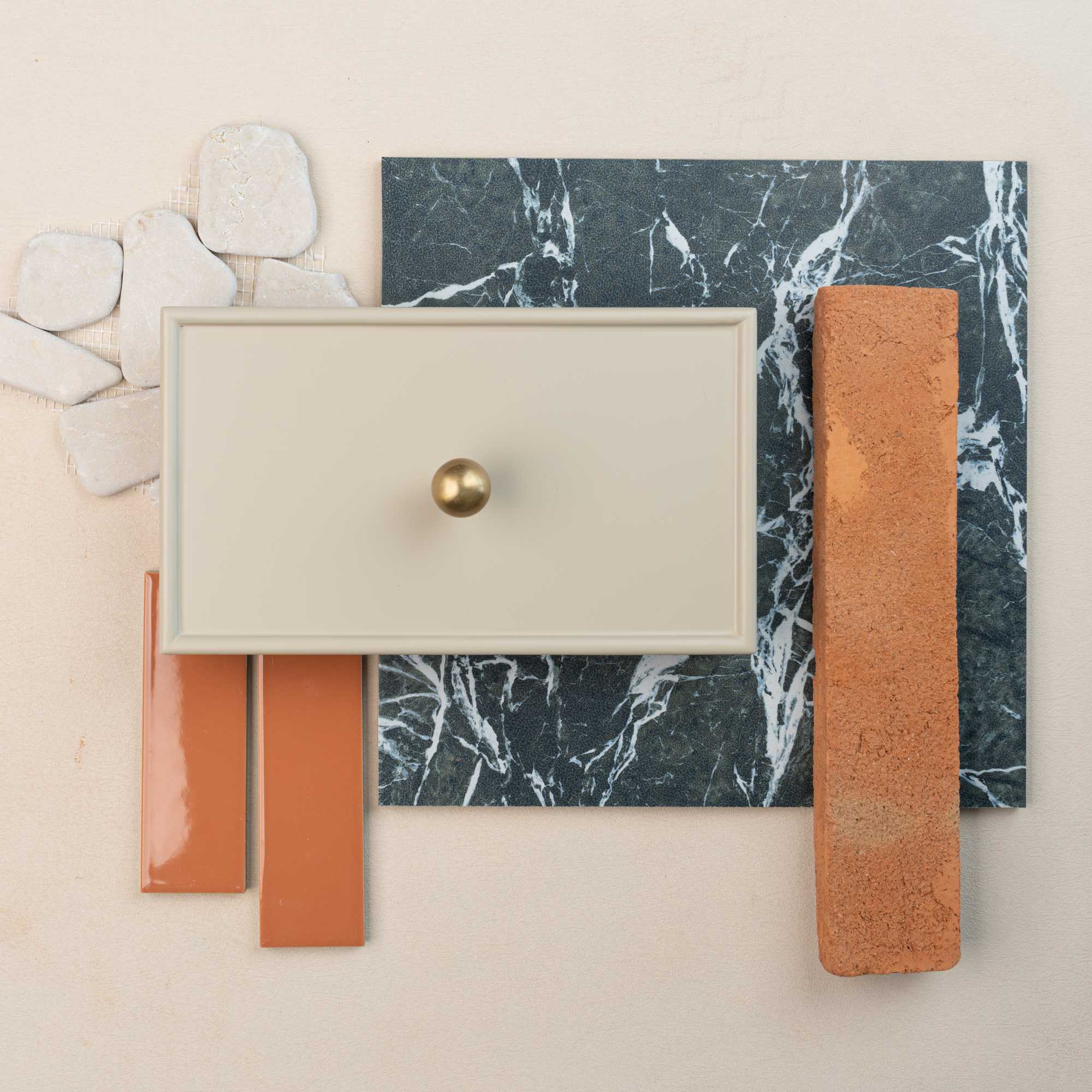 Flat lay of black marble tile, beige cabinet drawer front with brass knob, terracotta brick, orange ceramic tiles, and white stone pieces on beige background