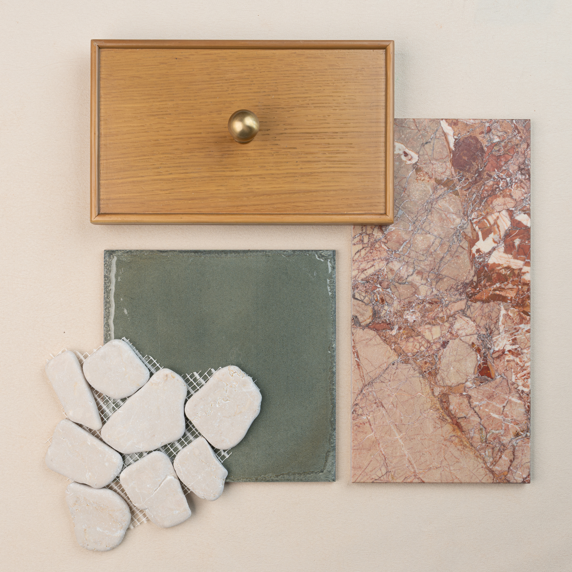 Flat lay of light oak cabinet drawer front with brass knob, green ceramic tile, pink marble tile, and white stone mosaic pieces on neutral surface