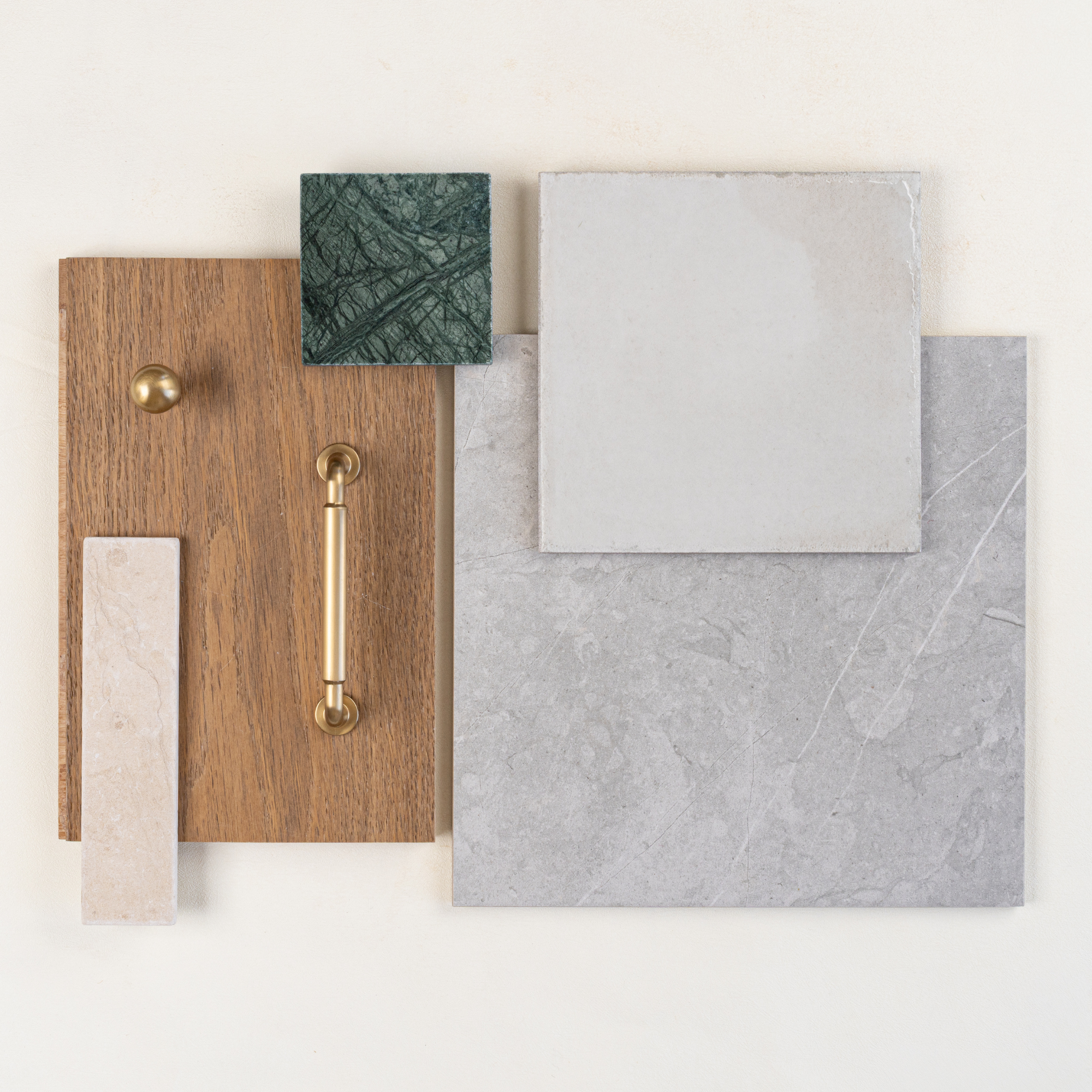 Flat lay of neutral stone tiles, green marble sample, oak wood with brass handle, and beige ceramic tile on pale background