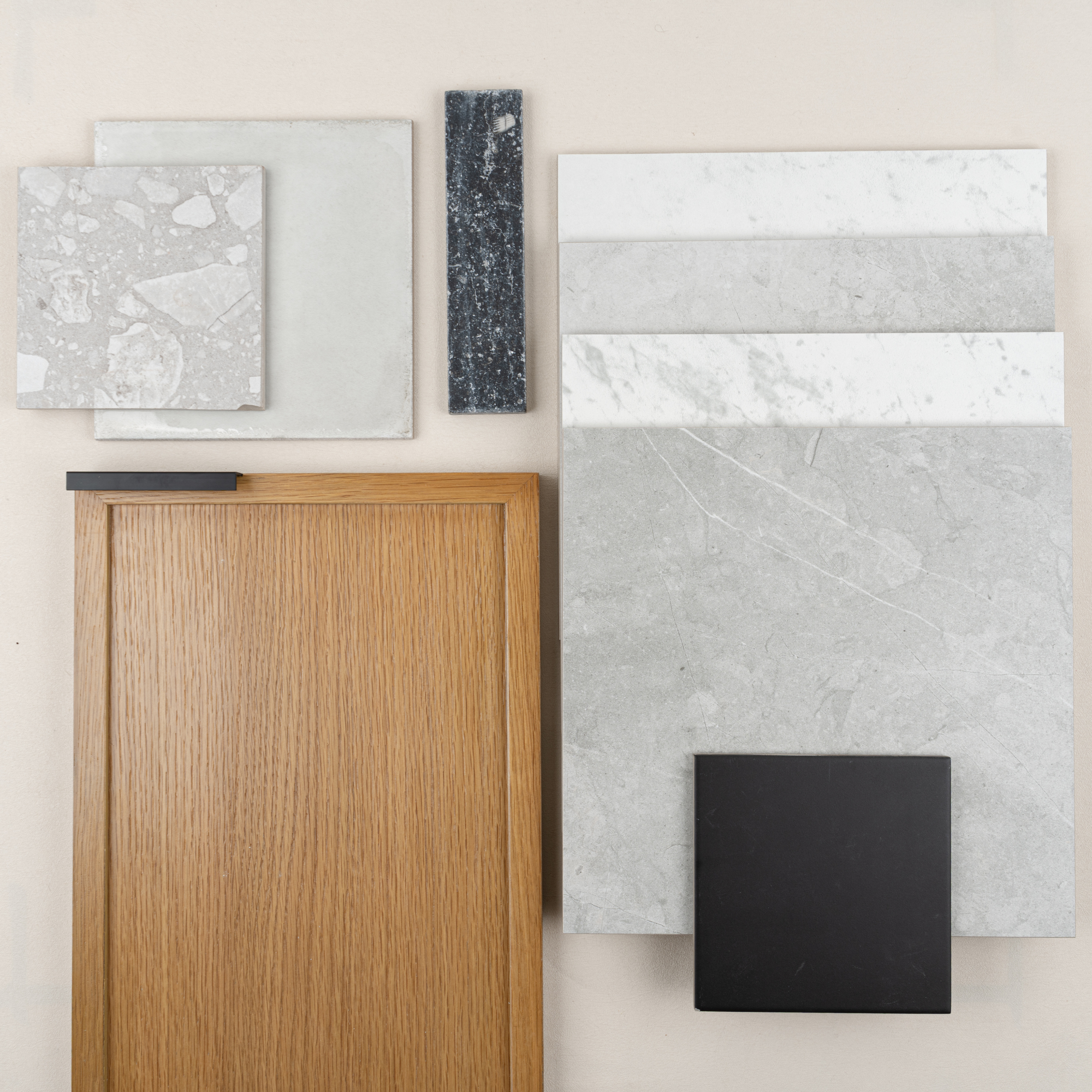 Selection of grey and white stone tile samples alongside an oak wood cabinet door and black hardware piece on a light surface